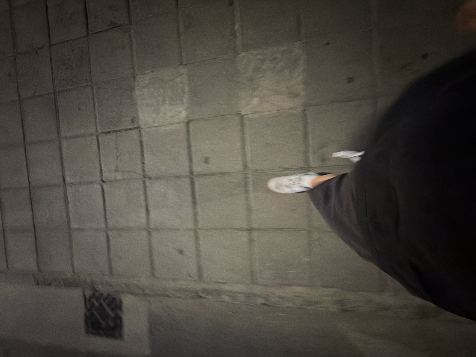 The image shows a first-person perspective of someone walking on a tiled sidewalk in Agios Athanasios Municipality, Cyprus. The person is wearing dark clothing and white sneakers. A drain grate is visible on the left side of the frame. The lighting suggests it is nighttime or a dimly lit area. The focus is on the ground and the lower part of the person's body, indicating a motion blur consistent with walking.