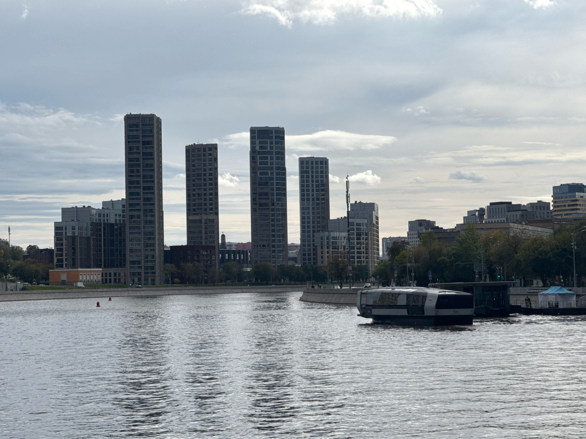This image captures a panoramic view of a cityscape along a wide river under a cloudy sky. The Moscow River is in the foreground, reflecting the muted light. On the far bank, a line of modern, tall buildings stands out, characterized by their sleek, geometric designs and numerous windows. These skyscrapers create a distinct urban silhouette against the sky.
In the middle ground, the riverbank is lined with trees, suggesting a green buffer between the water and the buildings. A paved promenade or walkway runs along the river, with some scattered figures visible, indicating a public space. A contemporary boat or ferry with a glass-enclosed cabin is moored or moving in the river, positioned to the right of the frame. It appears to be a passenger vessel.
Further along the riverbank, other structures are visible, including what appears to be a contemporary pavilion or cafe. Two red buoys are positioned in the river, marking a channel or hazard. The overall scene is one of a modern urban environment integrated with a significant waterway. The sky is overcast with scattered clouds, casting a diffused light on the scene. The architecture suggests a relatively modern development in Moscow, Russia.