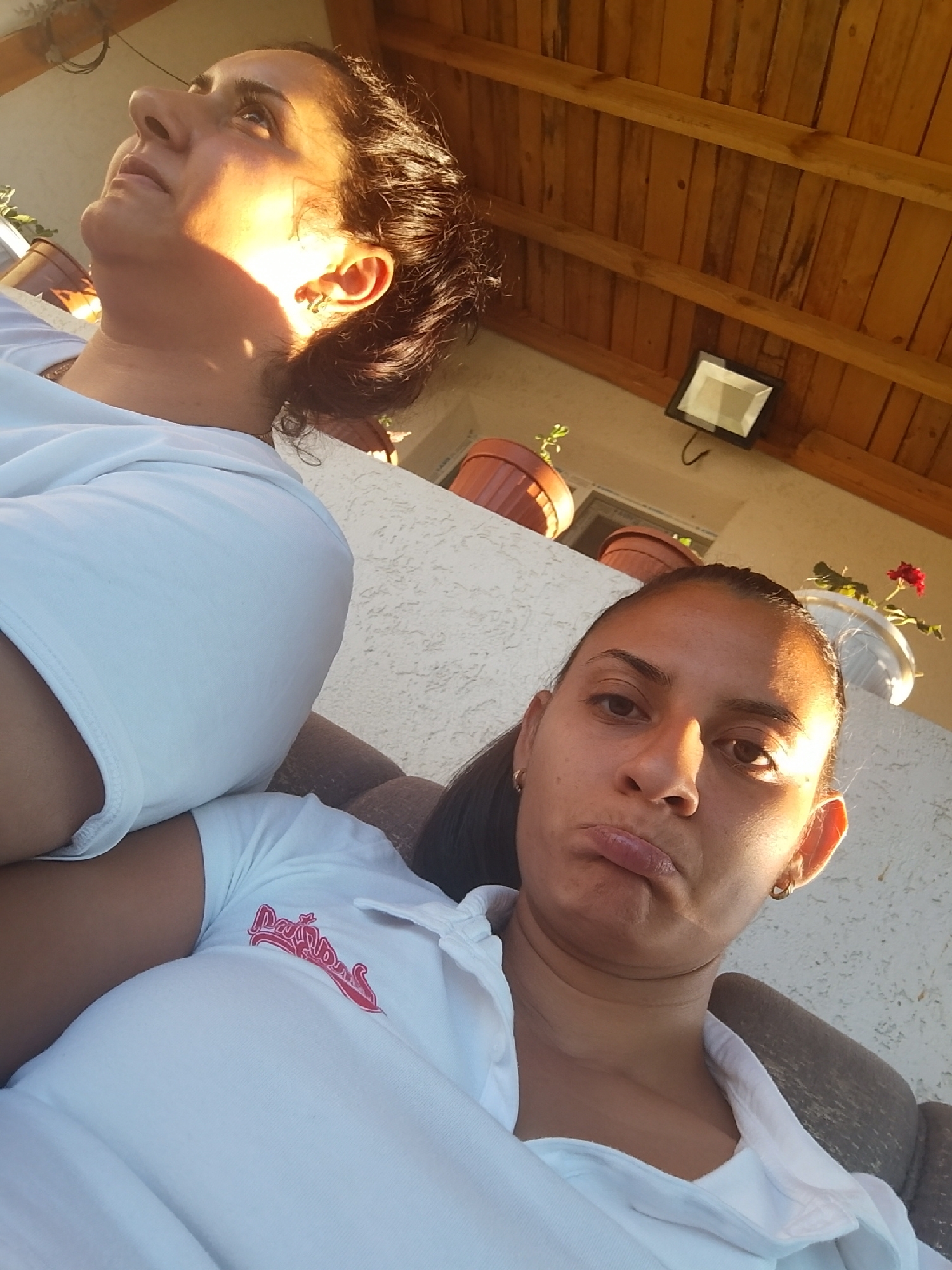 Two women are posed for a selfie outdoors. The woman in the foreground is facing the camera with her lips pursed. She is wearing a white polo shirt with a red logo. The woman in the background is looking upwards and to the left. She is wearing a white t-shirt. The background shows a wooden ceiling, a light fixture, and potted plants on a ledge. The scene appears to be in Zimnitsa, Bulgaria.
