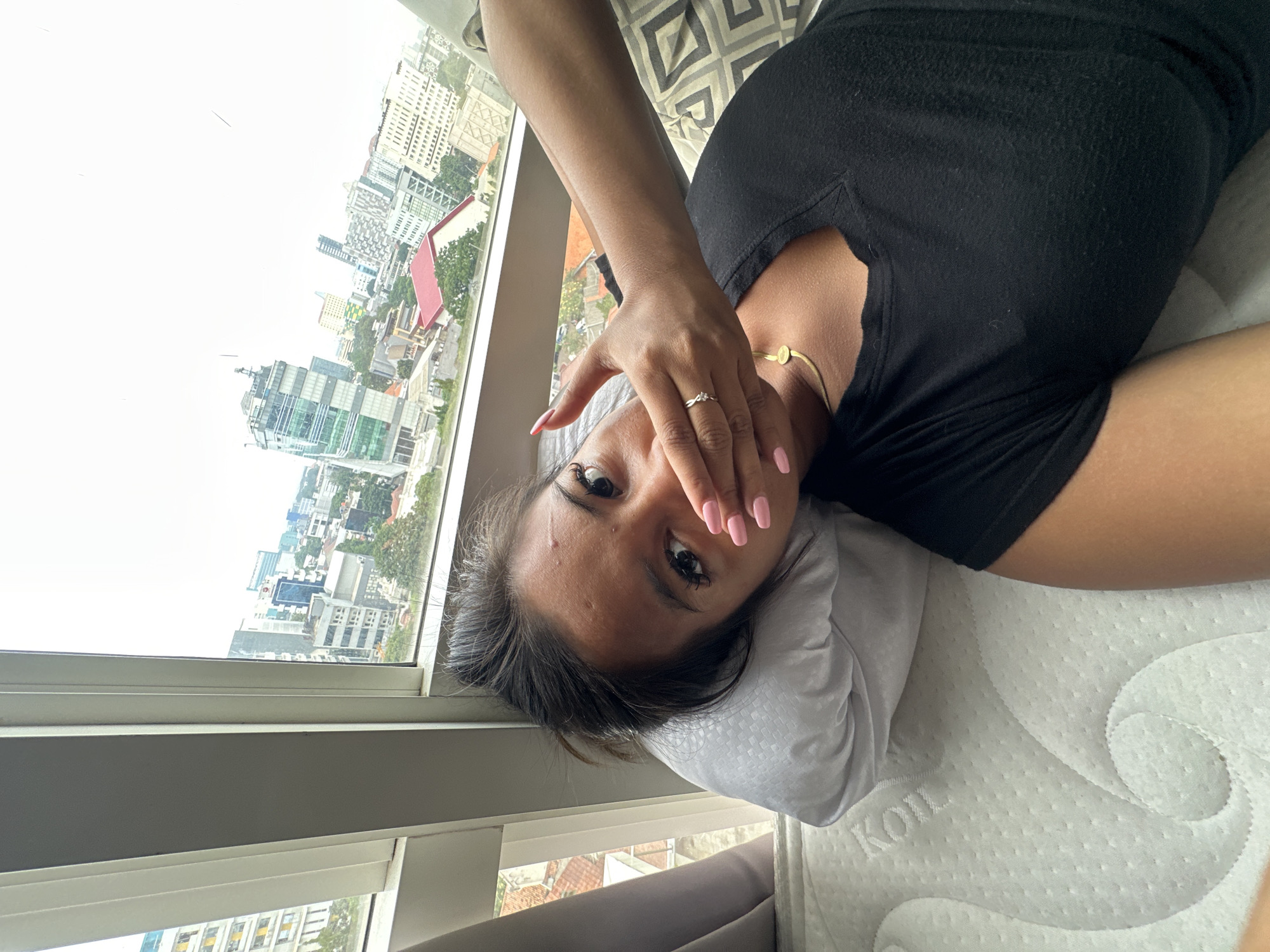 A woman lies on a white mattress near a window with a view of the city of Central Jakarta, Indonesia. The woman has dark hair and is wearing a black shirt. She covers her mouth with her right hand, on which she wears a ring, and looks at the camera. A gold necklace is visible around her neck. The window shows a cityscape with numerous buildings.