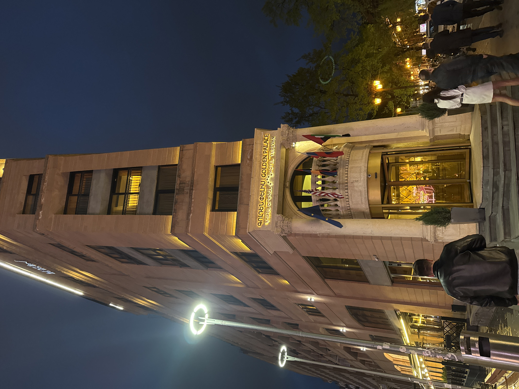 This image depicts a vibrant nighttime scene on a street in Yerevan, Armenia, focused on the brightly lit facade of a hotel. The building, constructed from light-colored stone or concrete, features multiple stories with numerous rectangular windows, some revealing interior lighting.
Prominently displayed above the main entrance is a gold-lettered sign in both Armenian and English, which reads "ԳՈԼԴԵՆ ՊԱԼԱՍ GOLDEN PALACE HOTEL YEREVAN." Below this sign, a series of international flags are hung, including the United States flag, above the hotel's revolving glass entrance doors.
The scene takes place at night, under a dark blue sky. The building's facade is illuminated with warm, golden lights, and several modern, ring-shaped streetlights cast a bright glow on the street and sidewalk.
Several people are visible. Near the hotel entrance, a group of individuals is gathered, some appearing to ascend steps. In the immediate foreground, a person is seen from behind, wearing a black leather jacket. Further down the street, more figures are discernible under the distant streetlights. Lush green trees line the street, with a faint, circular light pattern visible on some leaves, suggesting decorative lighting. The overall atmosphere is that of a lively urban evening.