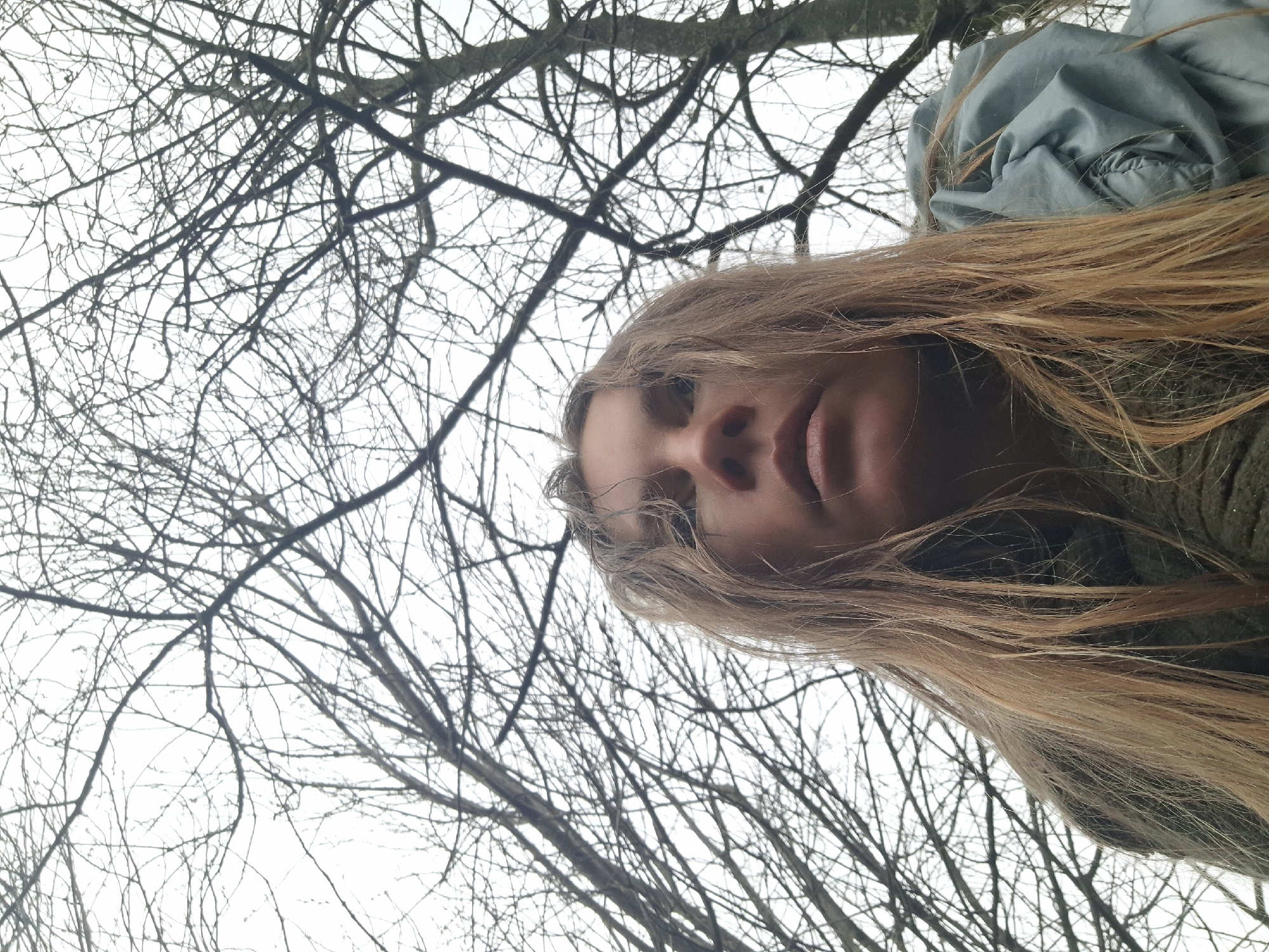 The image is an upward-facing shot of a person with long, blonde hair. Only the lower portion of the face, including the nose and mouth, is visible. The person is wearing a green shirt. Behind the person, filling the majority of the frame, is a dense network of bare tree branches against a grey sky. Part of a light blue jacket is visible in the top right corner. There is no information in the image to locate the scene in Khlevnoye, Russia, or any city specifically.