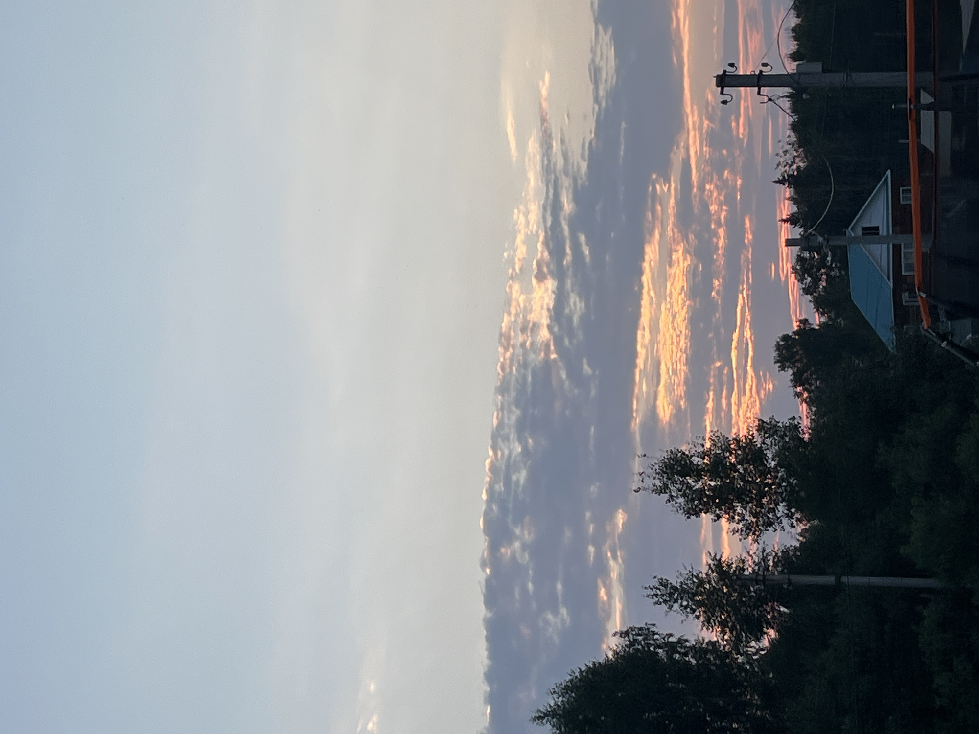 This is a photograph of a sunset over a rural landscape, likely in or near the city of Chaikovsky, Russia. The sky is filled with dramatic clouds, illuminated by the setting sun with hues of orange, pink, and yellow. Silhouetted against the vibrant sky are dark green trees and a small house with a bright blue roof. A utility pole with visible wires stands prominently in the foreground. The overall scene suggests a peaceful, twilight setting in a natural environment.