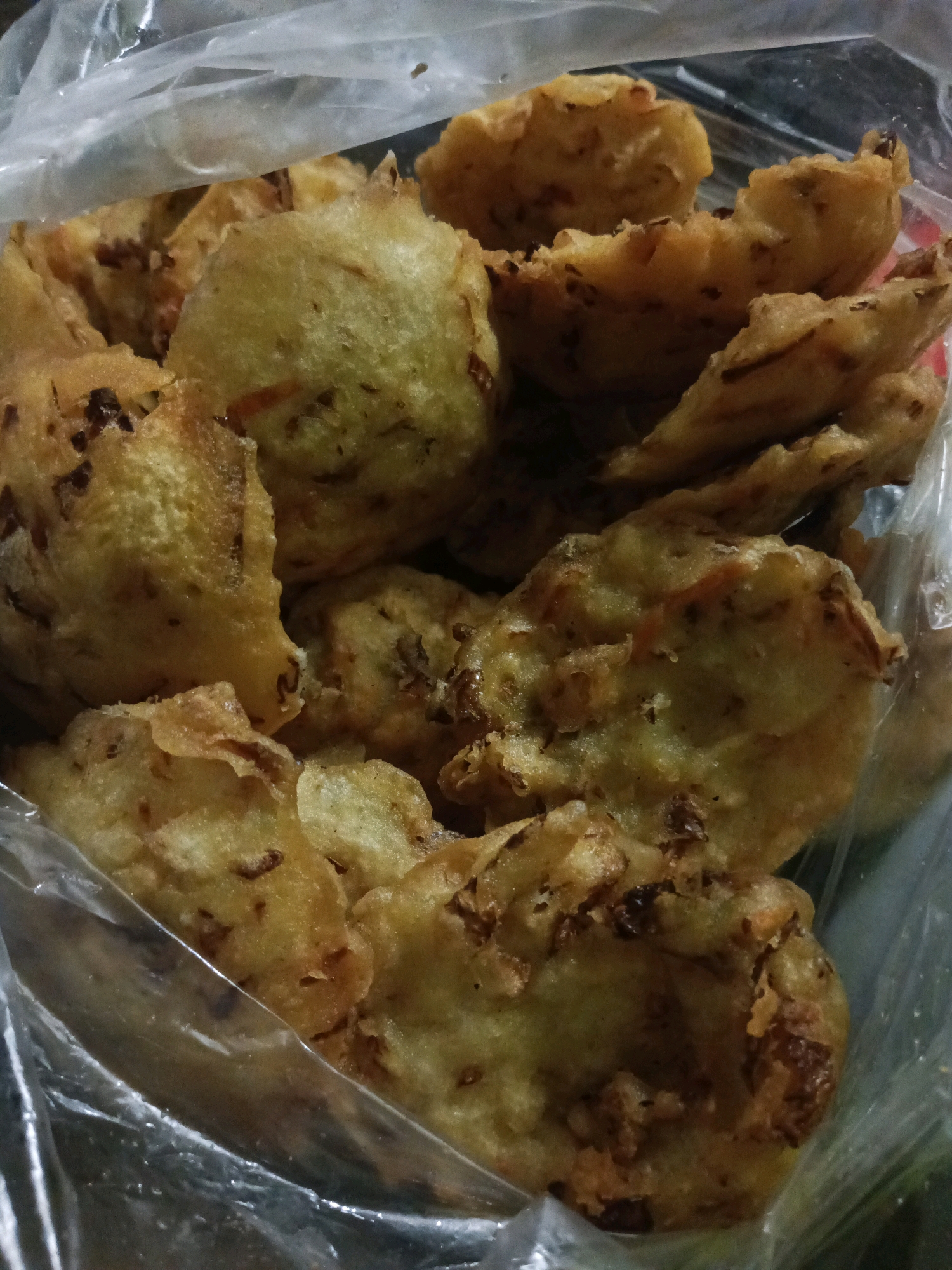 A close-up, top-down shot shows a pile of fried food, likely fritters, in a plastic bag. The fritters are golden brown and appear to be made with shredded vegetables, possibly carrots and onions, mixed into a batter. The texture is irregular, with some pieces having crispy edges and others being softer. The food is illuminated by overhead light, creating some glare on the plastic bag. The background is dark and indistinct, with the focus solely on the food items. There are no people or other objects visible in the image. This image was taken in Ngrendeng, Indonesia.
