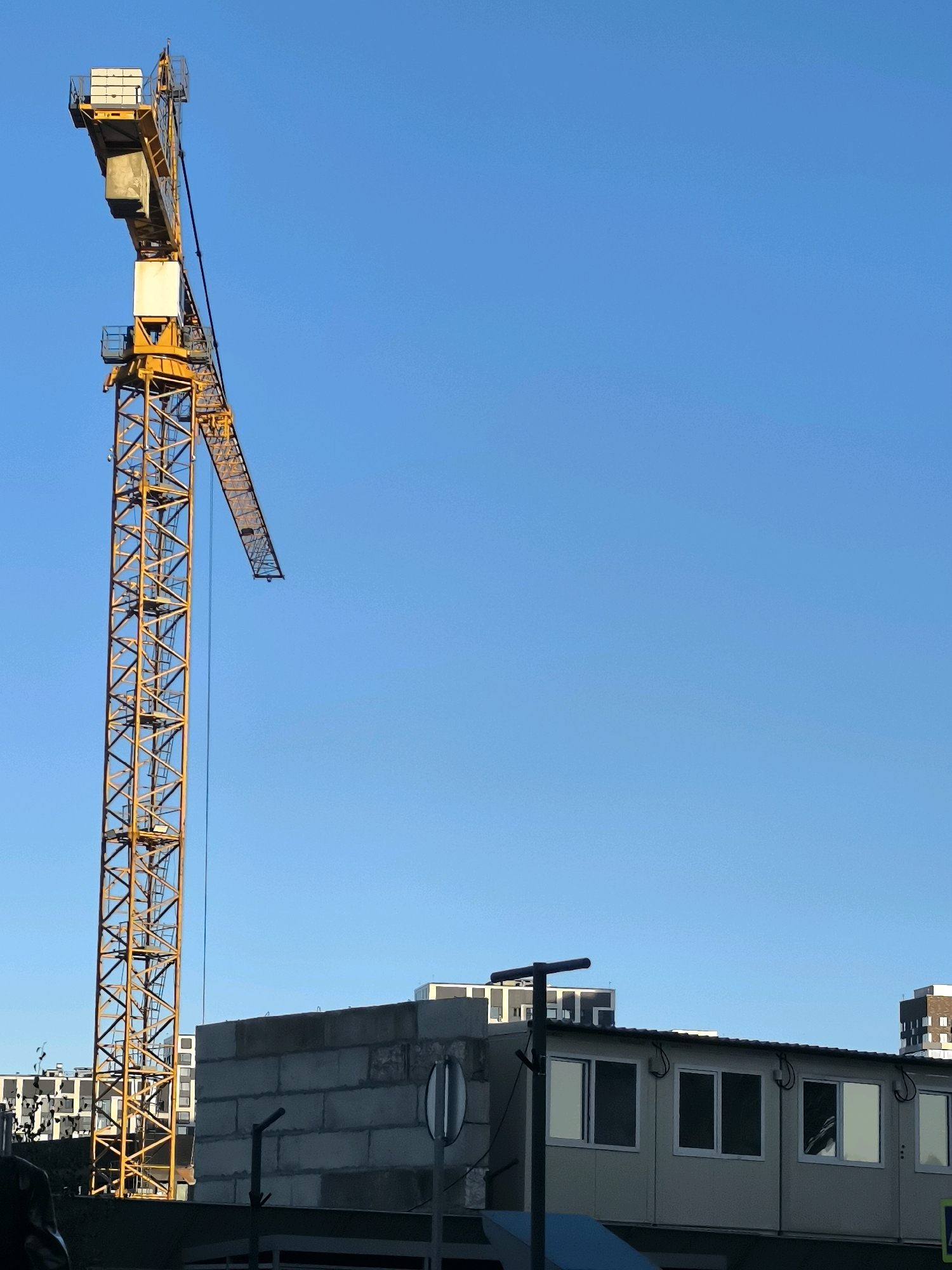 In Khimki, Russia, a towering yellow construction crane dominates the left side of the frame against a clear blue sky. The crane's jib extends diagonally towards the right. In the mid-ground, a low-rise building, likely a temporary site office or modular structure, is visible, featuring several windows. Behind this structure and further to the left, the facades of apartment buildings can be seen. A street sign with a circular white face and vertical black stripes is positioned in front of the modular building. The overall scene depicts an active construction site under a bright, cloudless sky.