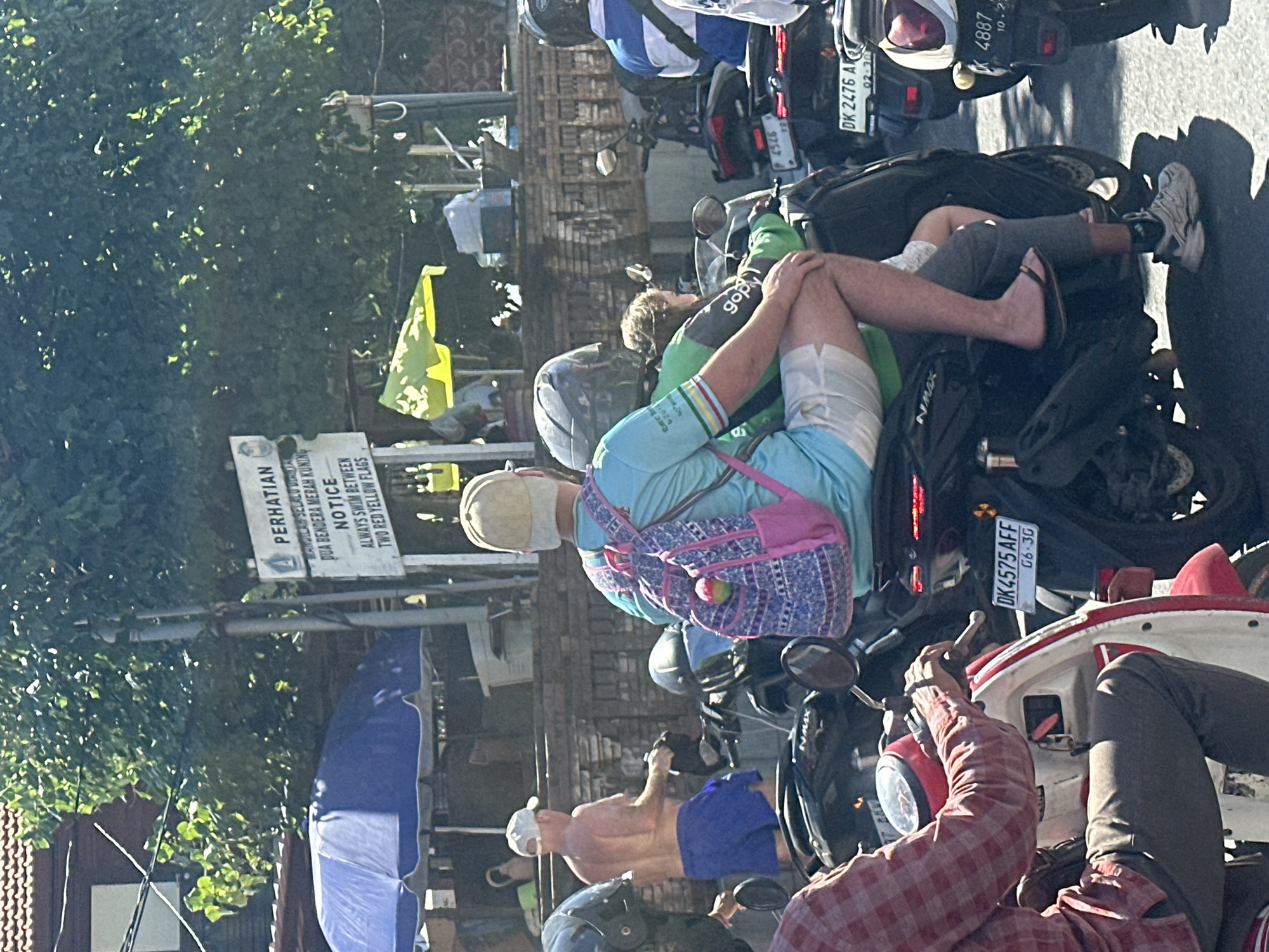 In Kuta, Indonesia, a street scene unfolds under bright sunlight. A group of people on motorcycles navigate the bustling street. In the foreground, a person in a red and white checkered shirt is riding a white and red scooter. Behind them, two individuals are seated on a black motorcycle. The person in the front wears a light blue t-shirt, a beige cap, and a pink and blue patterned backpack. They are followed by a person in a green jacket, with the "gopay" logo visible on their sleeve. Further down the street, another person in a blue and white striped shirt is visible on a motorcycle. A sign in the background, in Indonesian and English, warns: "PERHATIAN MANDIILAH SELALU DIDALAM DUA BENDERA MERAH KUNING NOTICE ALWAYS SWIM BETWEEN TWO RED YELLOW FLAGS." The environment suggests a tropical locale, with lush green trees and buildings lining the street.