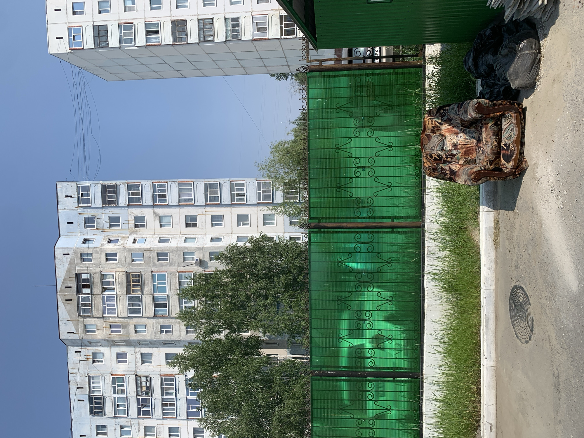 The image shows an outdoor scene in the city of Megion, Russia. In the foreground, a worn armchair with a floral pattern sits on a paved surface next to a pile of black trash bags. Behind the armchair is a tall, green, translucent fence with decorative black metalwork. Beyond the fence, a strip of green grass and trees is visible, separating the fence from two tall, multi-story apartment buildings. The buildings are constructed from light-colored concrete and have numerous windows. The sky is clear and blue. The scene suggests an urban environment, possibly an alley or courtyard area, where discarded items are present.