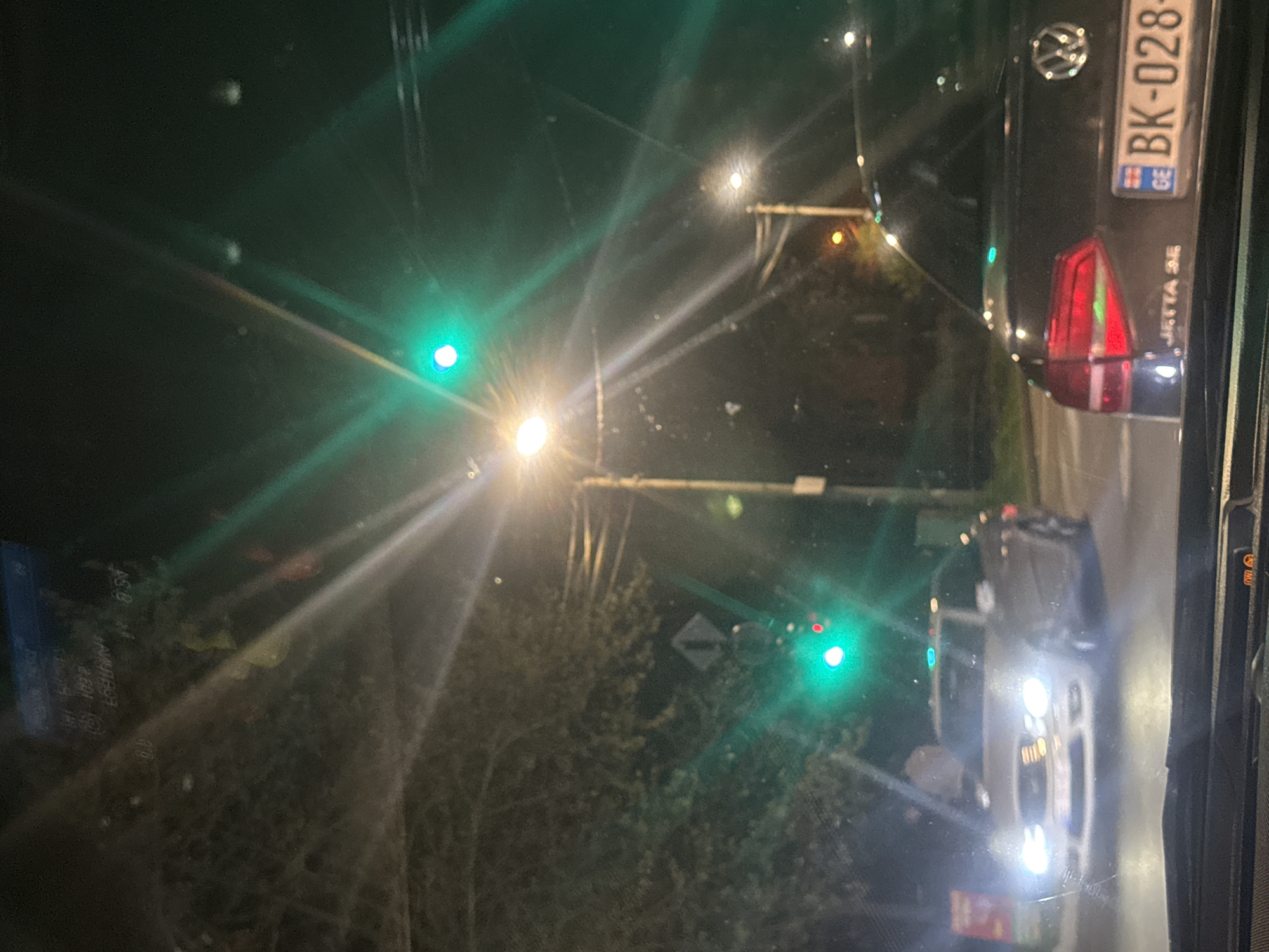 The image depicts a night scene in Tbilisi, Georgia, showing traffic lights, vehicles, and streetlights. A black Volkswagen with license plate BK-028-GE is positioned ahead and to the right. Another vehicle with headlights on is visible further down the road. The traffic lights display a green signal. The location is implied by the Georgian license plate. The environment is urban, with streetlights and surrounding vegetation.