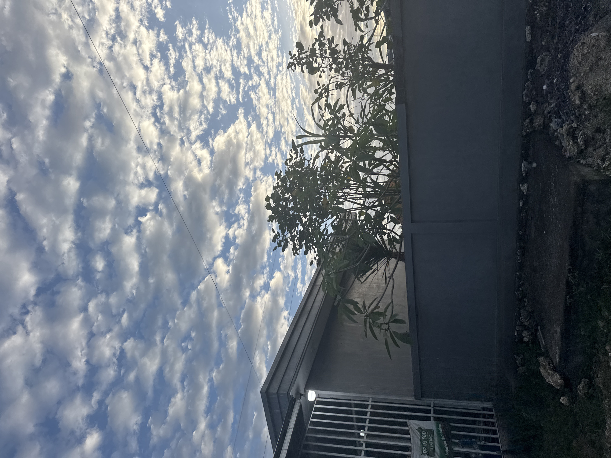 The image shows an outdoor scene with a fence and trees in the foreground, and a cloudy sky in the background. The fence is dark gray and made of concrete. Behind the fence, there are several trees with green leaves. Above the trees, the sky is filled with white and gray clouds, with patches of blue sky visible. There is a building to the left of the image, with a light on its exterior. A power line stretches across the top of the frame. The ground in the foreground is covered with dirt and rocks. The lighting suggests it is either early morning or late afternoon.
