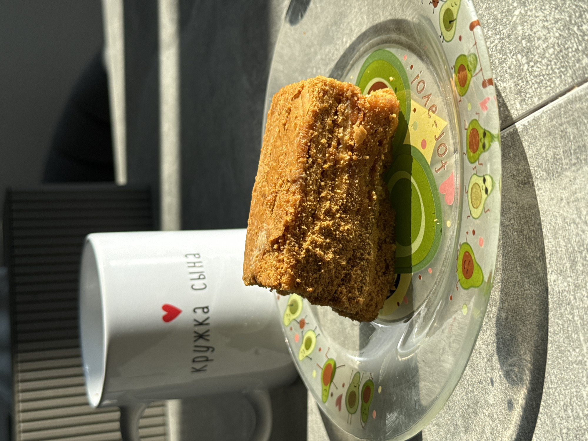 A slice of honey cake rests on a clear glass plate with an avocado pattern. To the left of the plate is a white mug with a red heart and Russian text that reads "кружка сына" (mug for son). The scene is set on a gray, tiled surface, bathed in sunlight that casts long shadows. The background shows a muted, textured object, possibly a radiator cover. The overall setting appears to be an indoor domestic environment, likely a kitchen or dining area. No people are visible. The image has a warm, slightly overcast lighting.