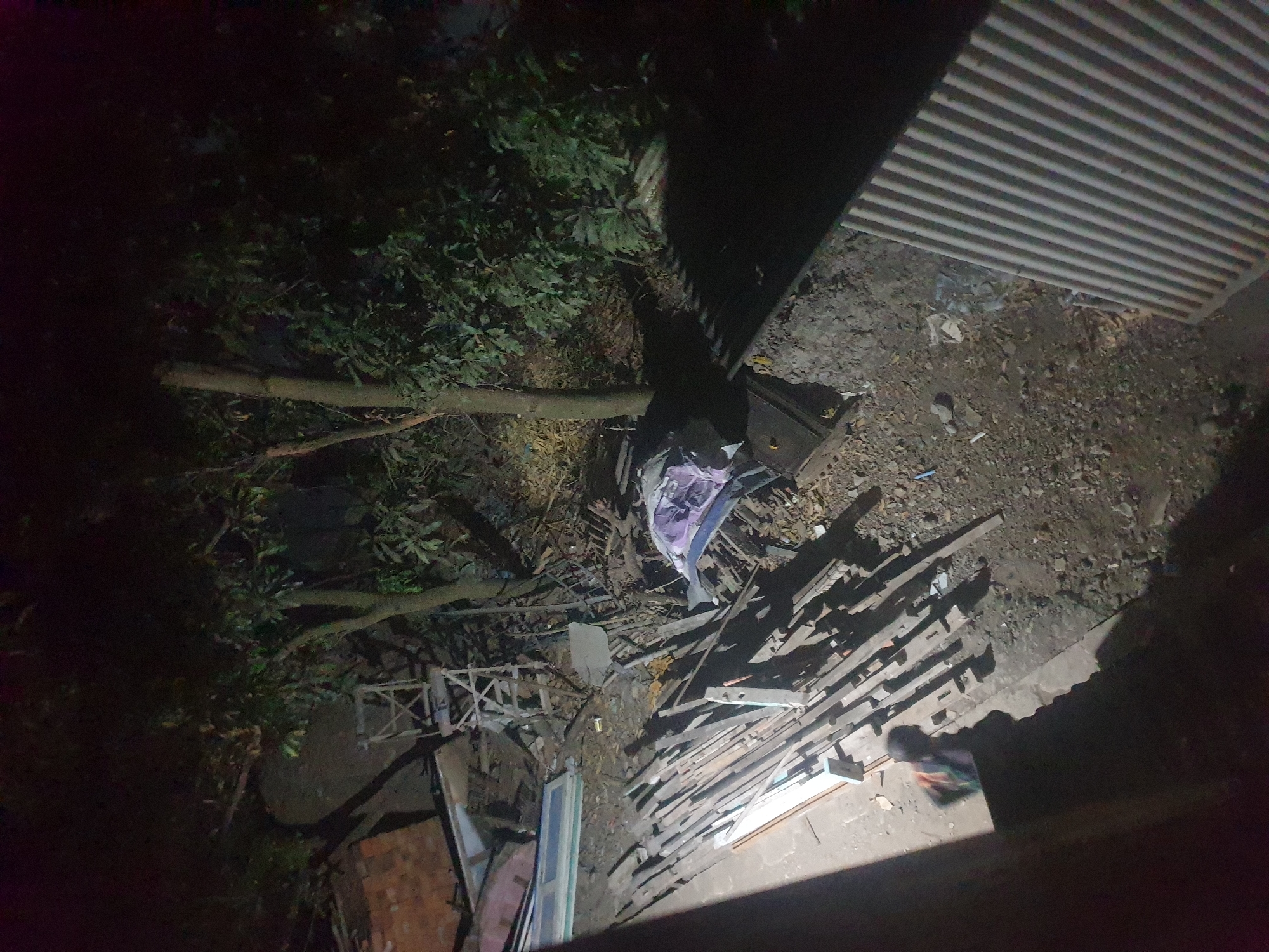 The image depicts an outdoor scene at night in what is identified as the city of Keboharan, Indonesia. The environment appears to be a cluttered, unkempt area with piles of building materials, including long wooden planks and corrugated metal sheeting. Vegetation, including trees with dense foliage, is visible in the background. A dark, indistinct figure, possibly a person, is partially visible in the foreground, casting a shadow. Scattered debris litters the ground. A small pile of fabric, appearing to be a purple and white blanket or cloth, lies on the ground. A ladder-like structure is also present among the debris. The overall impression is one of disarray and neglect.