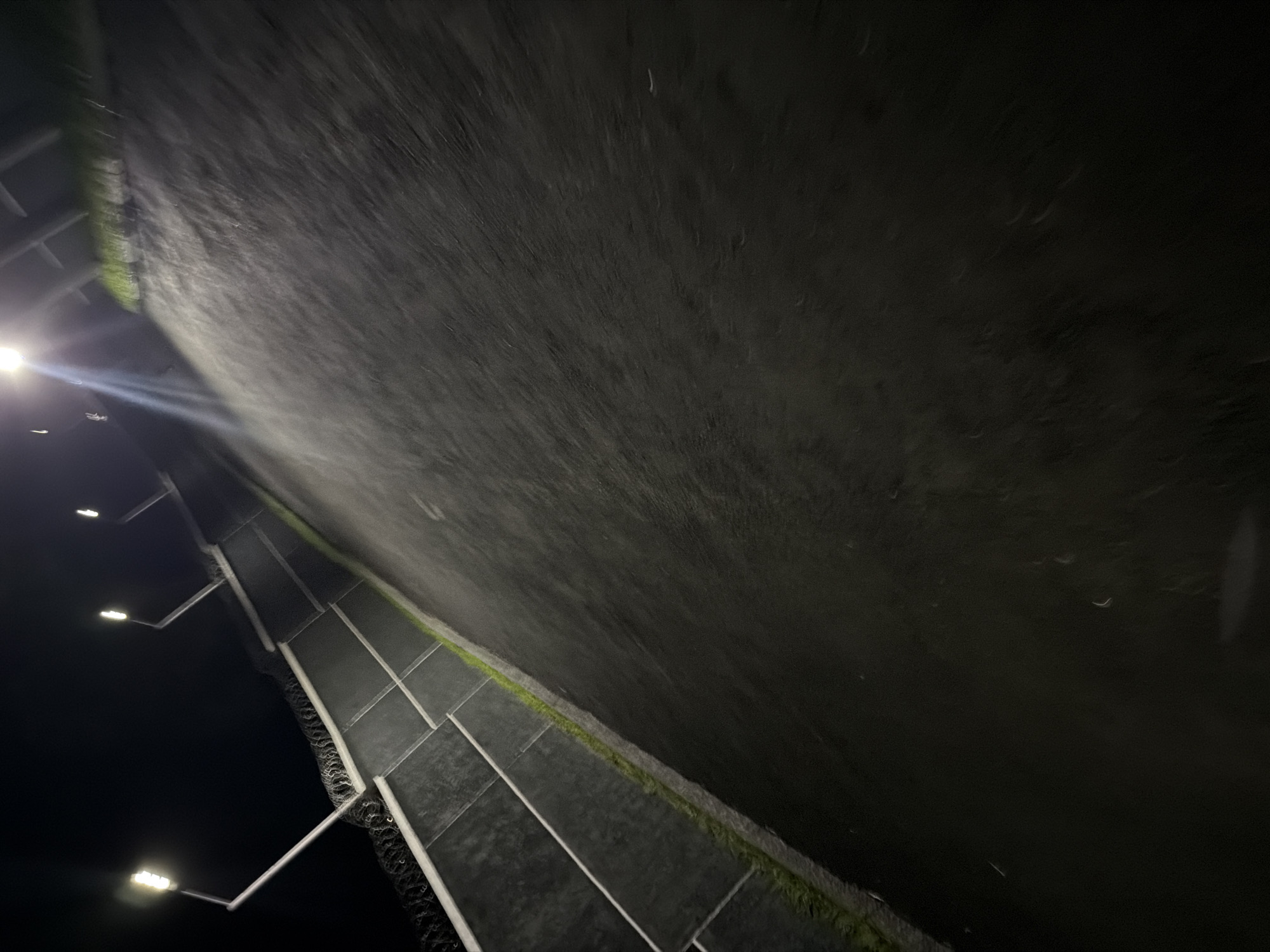 The image depicts an outdoor scene at night, illuminated by streetlights. A paved surface, possibly a road or pathway, occupies the majority of the frame, angled from the bottom left to the top right. To the left of the paved surface is a low, grey wall, segmented by lighter vertical lines. Along the top edge of this wall, razor wire is visible. A thin strip of grass or vegetation runs along the base of the wall, separating it from the paved area. Several streetlights are visible along the top of the wall, casting bright light downwards. The overall impression is that of a secured outdoor area, possibly a perimeter or walkway within a facility, at night. There are no people or animals visible in the image. The specific location in Bukuru, Nigeria, is not determinable from the visual information provided.