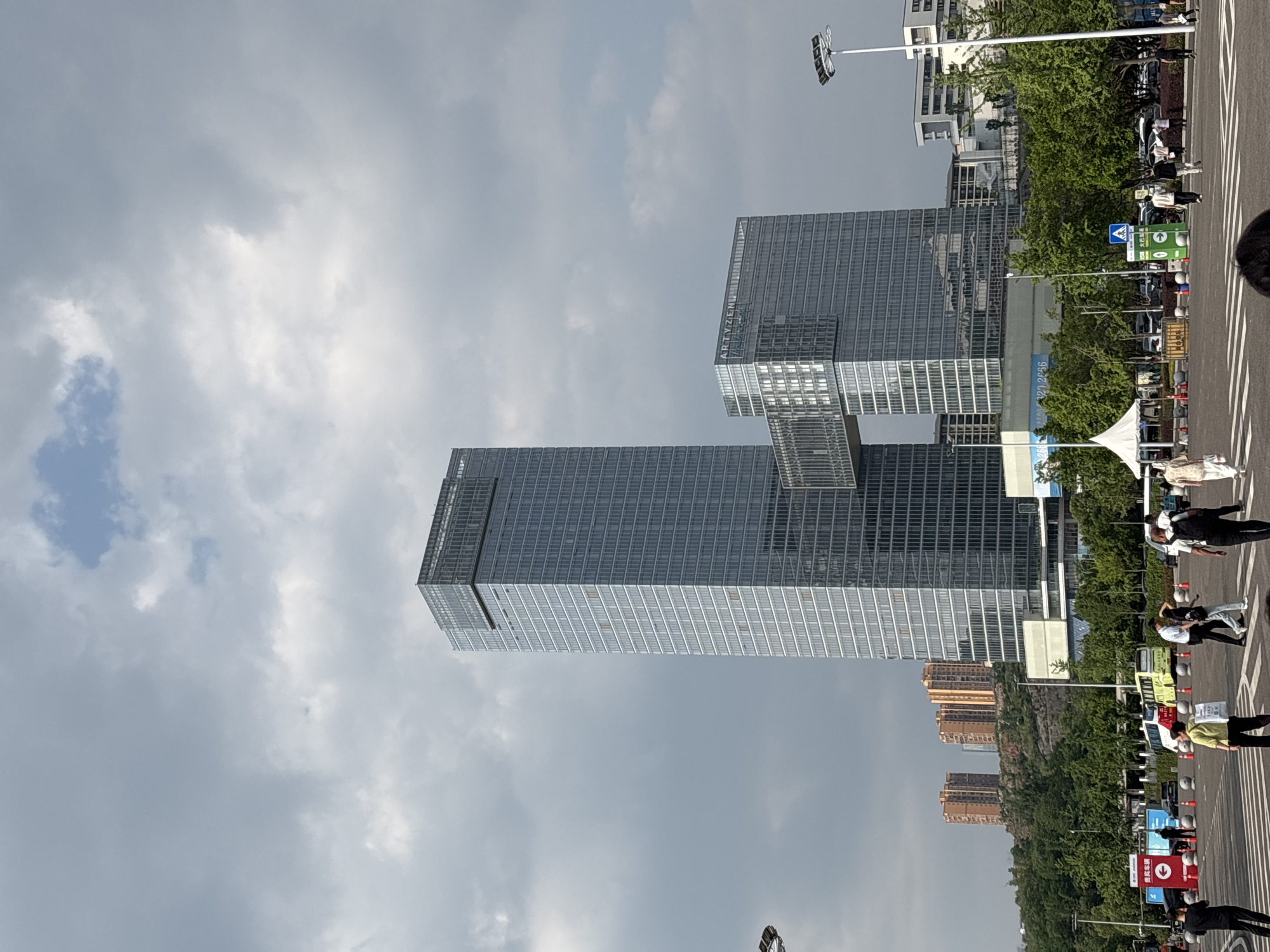 The image captures a street scene in Liangjiang, China, featuring two modern skyscrapers under a cloudy sky. The skyscrapers are characterized by their glass facades. Pedestrians walk on the street, which has white lane markings. Along the street, there are trees and various traffic or directional signs. In the background, one can see additional buildings and foliage, contributing to the urban setting.