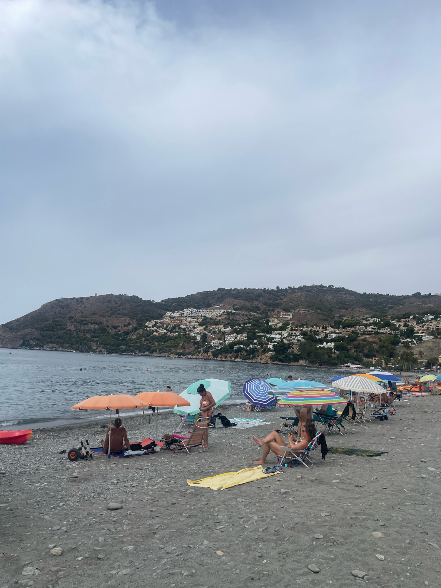 The image depicts a crowded beach in La Herradura, Spain, under an overcast sky. The beach is composed of dark pebbles and sand. Numerous beach umbrellas, in various colors and patterns, are erected, providing shade for beachgoers. People are seated on beach chairs, towels, and directly on the pebbles, some appearing relaxed and others engaged in conversation. A calm sea stretches towards a hillside covered with a town, characterized by white buildings, and lush vegetation. The distant hillside is crowned by a watchtower. Beach equipment, including chairs, towels, and a red kayak, are visible scattered across the shore. A few individuals are visible in the water.