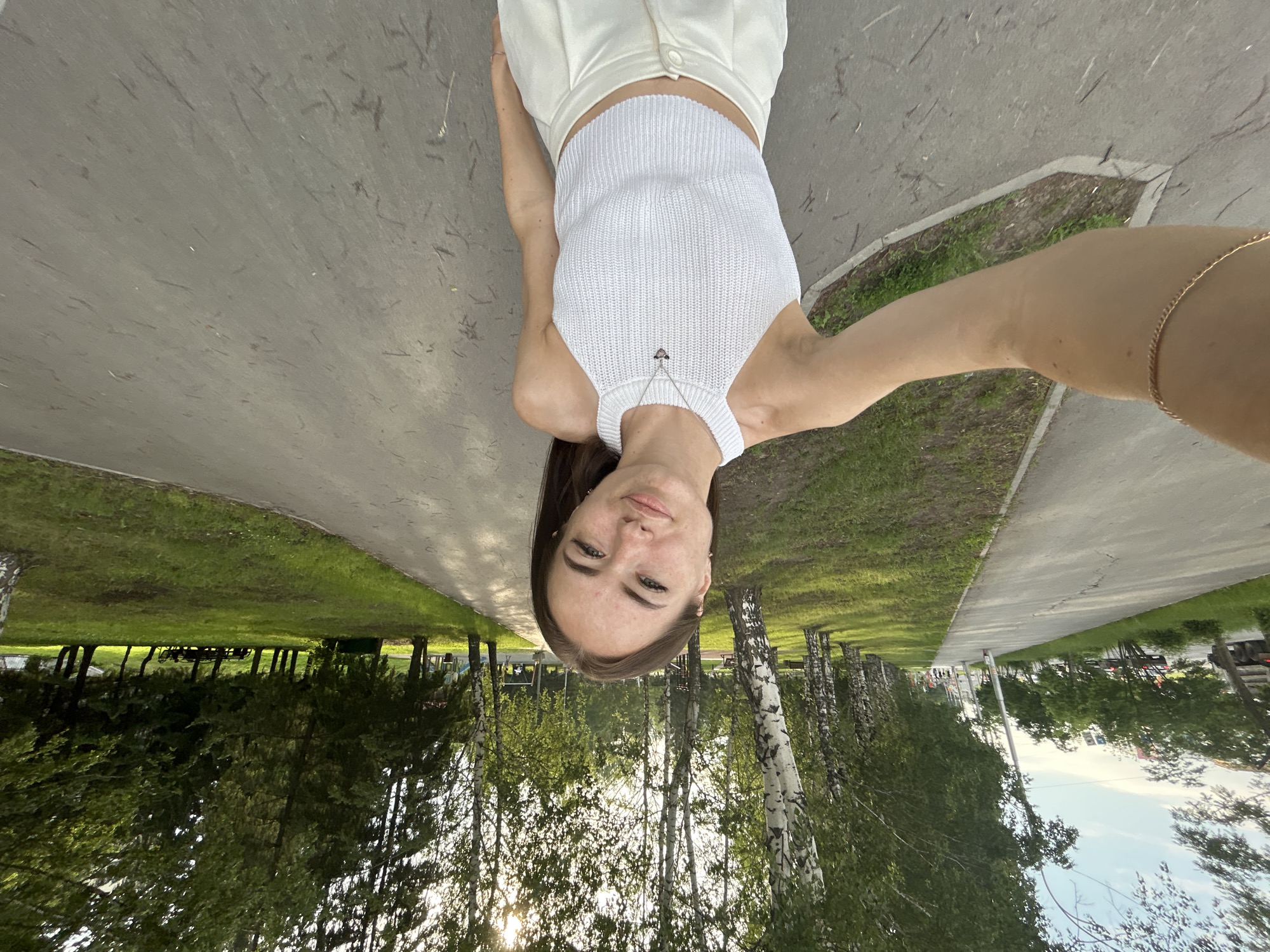 A young woman is taking a selfie outdoors in a park-like setting with a paved path. She is wearing a white halter top and white pants, and a silver necklace with a pendant. The path is bordered by a grassy area and a line of trees. In the background, there are more trees, grass, and a glimpse of buildings and cars, suggesting an urban environment. The sun is visible through the trees, casting a warm light. The woman's expression is relaxed and she is looking directly at the camera. The perspective of the selfie suggests she is holding the camera at arm's length.