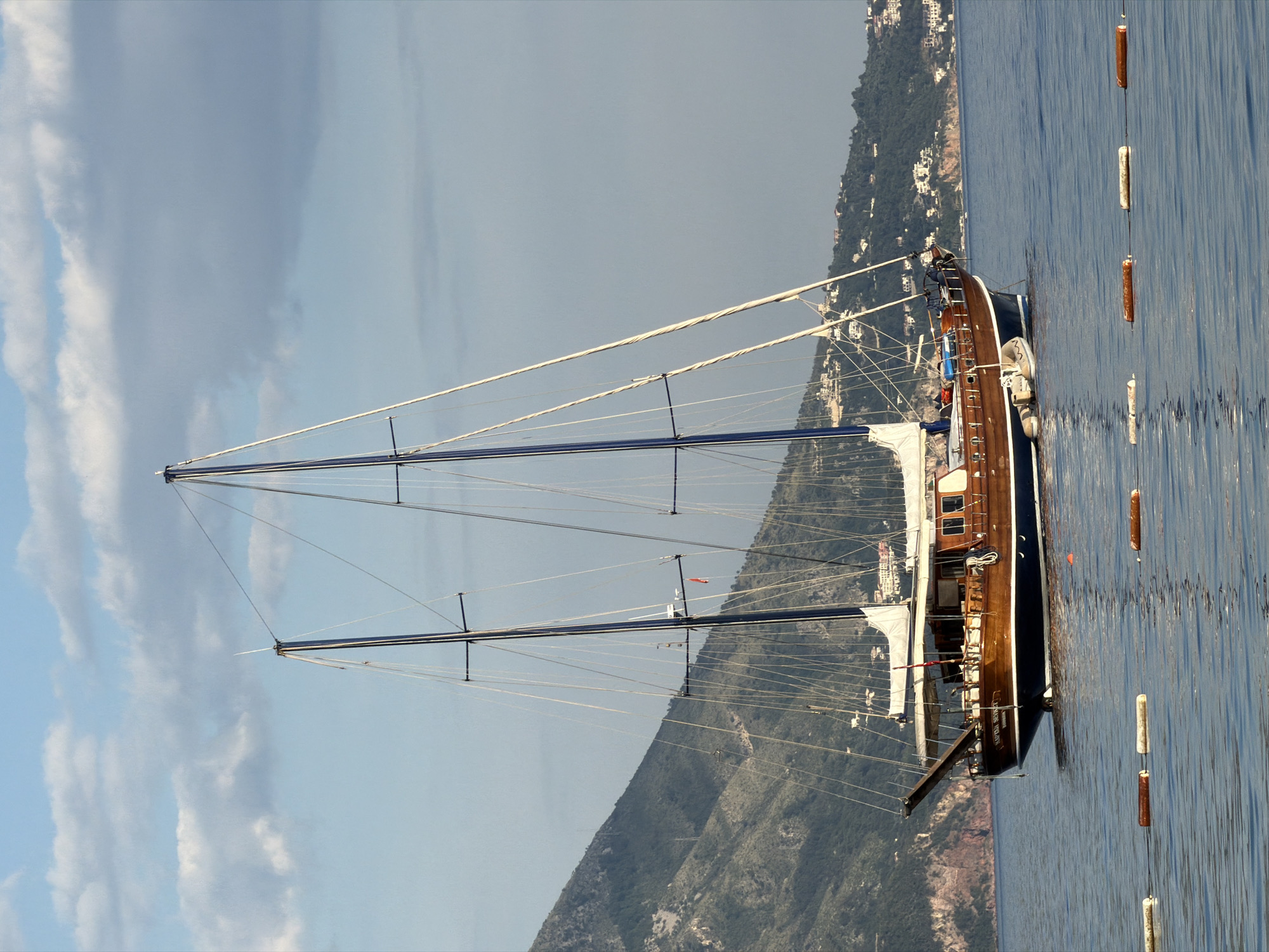 The image shows a sailboat in the Adriatic Sea, near the city of Budva, Montenegro. The sailboat, with two masts and several sails, is docked near a shoreline with mountainous terrain covered in vegetation and buildings. A string of buoys floats to the right of the boat. The sky above is overcast.