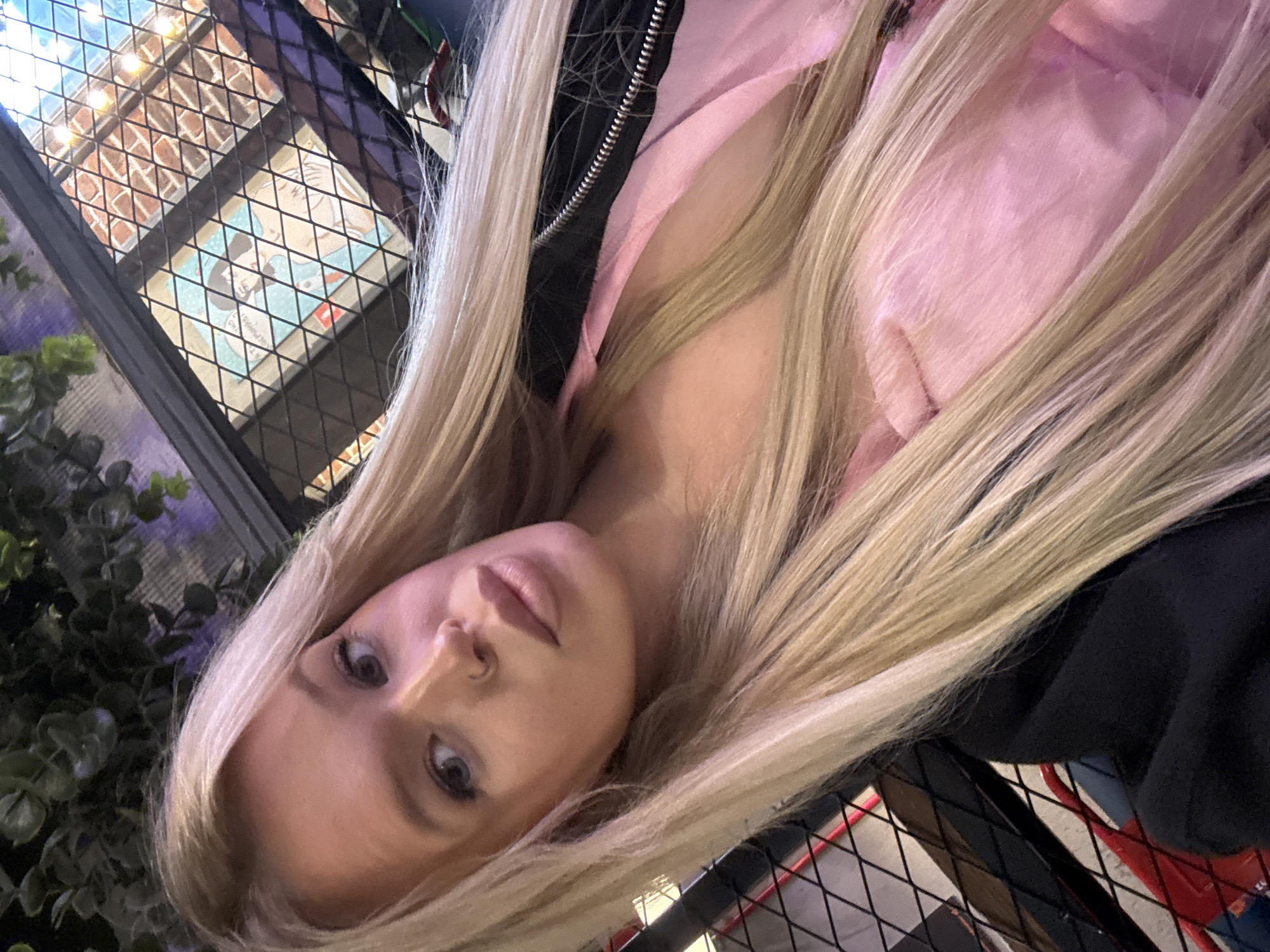 A blonde woman with long, straight hair is taking a selfie. She is wearing a pink top and a black jacket. She has a nose ring. The background is blurred, but appears to be an indoor setting with some greenery and a patterned window. The image is taken from a slightly high angle.