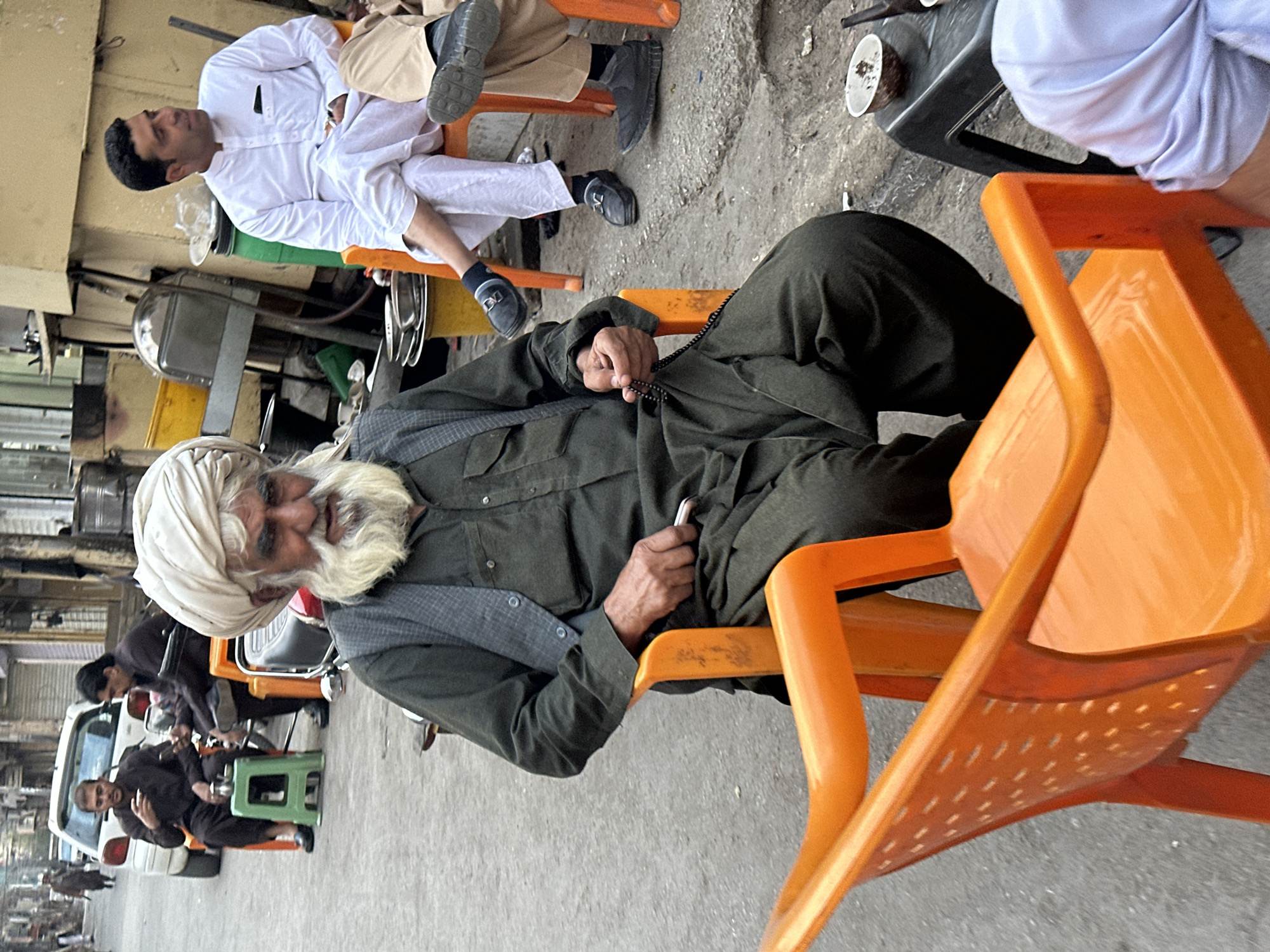 The image captures a street scene in Quetta City Tehsil, Pakistan. An elderly man with a white beard and turban is seated on an orange plastic chair, holding prayer beads in his hands. He is wearing a dark green outfit. Behind him, another man is sitting on an orange chair, dressed in white traditional clothing, with his legs crossed. Other individuals are seen in the background, some sitting on green plastic chairs. A white vehicle is partially visible on the left side of the image. The setting appears to be an outdoor gathering in an urban area, potentially a roadside establishment or meeting place. The street and surrounding buildings are visible in the background.