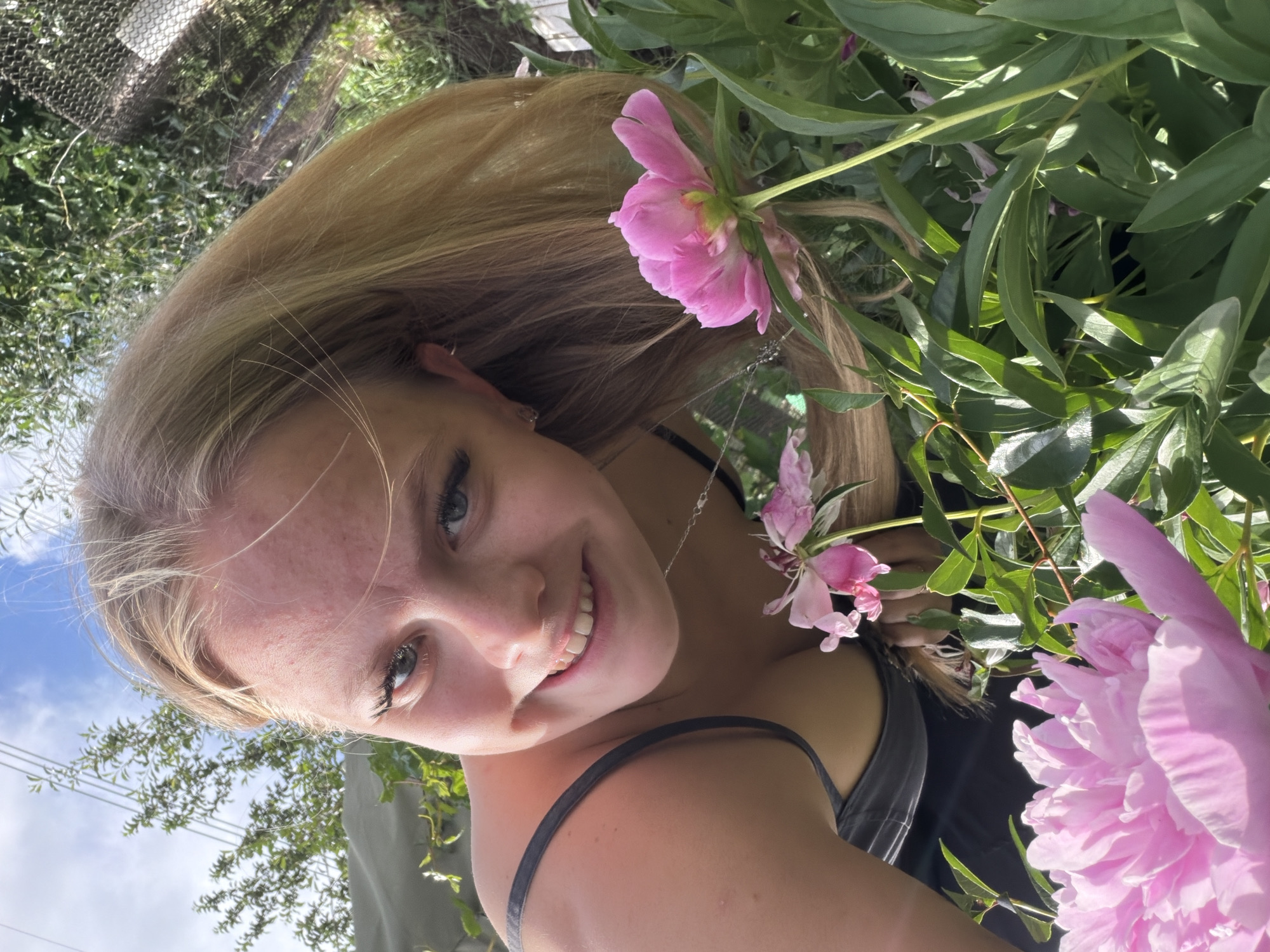 The media file is a portrait of a young woman with blonde hair, smiling and looking at the camera. She is outdoors, surrounded by lush green foliage and pink peonies. The sun is shining, casting dappled light on her face and the flowers, suggesting a warm, clear day. The background includes a glimpse of the sky and some trees, hinting at a natural or garden setting. The woman is wearing a dark tank top and a delicate silver necklace. Her expression is cheerful and relaxed. The image has been rotated, with the top of her head appearing to the left and her feet to the right. The overall mood is bright and summery, with a focus on natural beauty and youthful joy. No visible text is present in the image. The location context provided is Chaikovsky, Russia, which aligns with a likely outdoor setting in a temperate climate during blooming season.