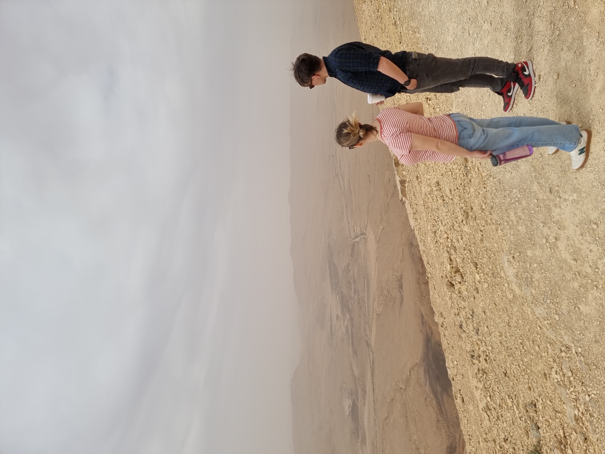 Two people stand on a desert overlook in Mitzpe Ramon, Israel. One person, wearing a blue plaid shirt, dark pants, and red/black sneakers, is next to a person with a pink and white striped shirt, blue jeans, and white sneakers. They are both looking out over the edge of a cliff at the desert landscape. The sky is overcast. A water bottle can be seen in the hands of the person with the striped shirt.