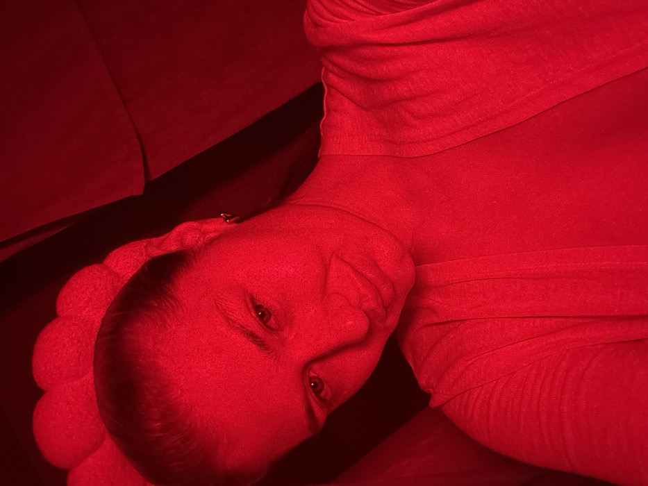 A person lies on their side, illuminated by a strong red light. The head rests on a pillow or cushioned surface. The face is visible, with eyes open and a slight smile. The person wears a cloth-like garment that drapes around the neck and chest. The red light casts a uniform hue over the subject and background, obscuring details and creating a vibrant, singular color palette. A dark area suggests furniture or a wall behind the person.