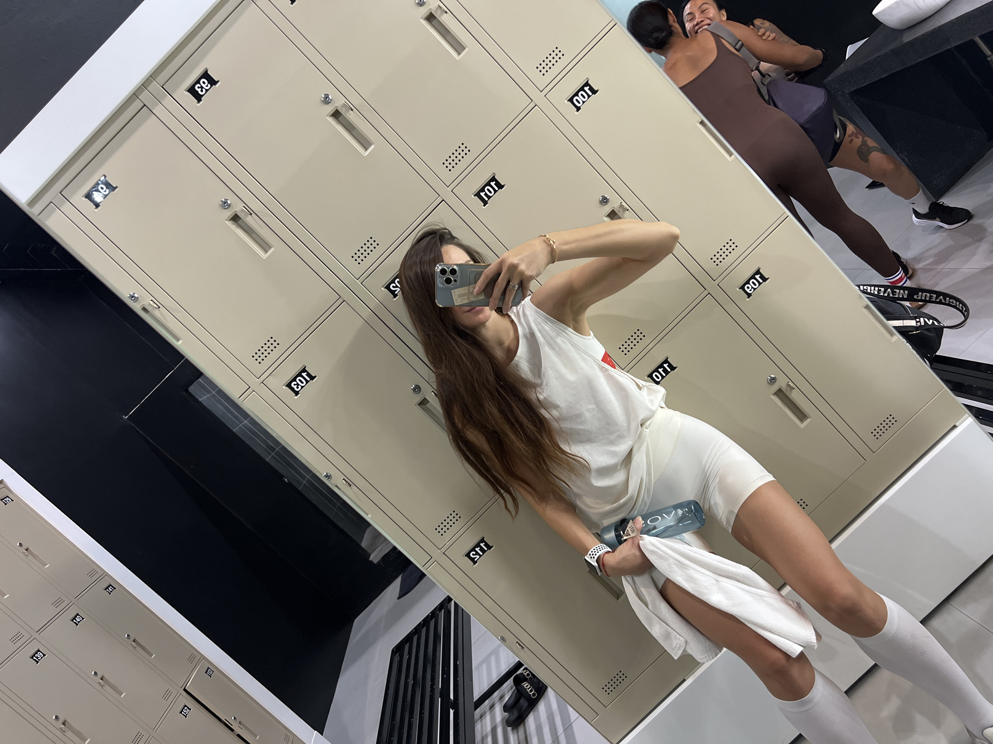 The image depicts a woman in a locker room, taking a selfie in front of a bank of lockers. She is wearing a white tank top, white shorts, and white socks. She is holding a water bottle and a white towel. There are two other women visible in the background, embracing. The locker room is brightly lit and appears to be clean. The city of Unknown, Unknown is not discernible from the image.