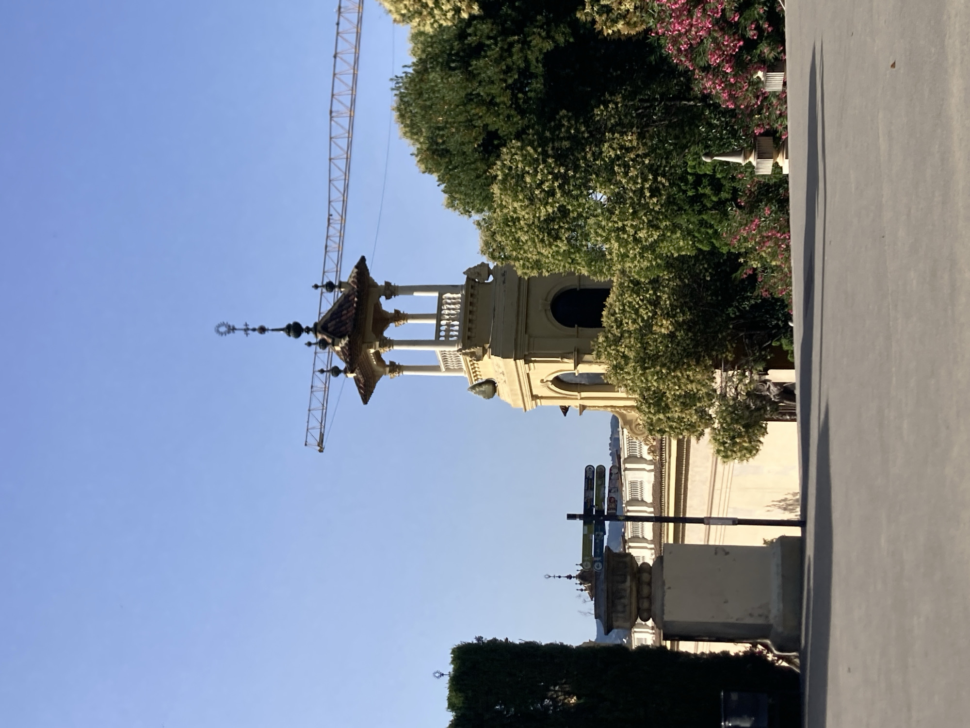 The image depicts a view towards a prominent yellow tower with ornate architectural details, including a decorative spire. A construction crane is visible in the background, extending across the clear blue sky. In the foreground, a paved pathway leads towards a landscaped area with lush green trees and vibrant pink flowers. A signpost with multiple directional signs stands near a stone pillar, and other architectural elements, such as ornate urns, are scattered throughout the scene. The overall impression is one of an outdoor public space, possibly a park or garden, in a city with a mix of historic architecture and ongoing development. The presence of Mediterranean flora suggests a warm climate. Based on the architectural style of the tower and the surrounding buildings, this scene is likely located in Barcelona, Spain, possibly within the Montjuïc hill area which features many such structures and gardens.