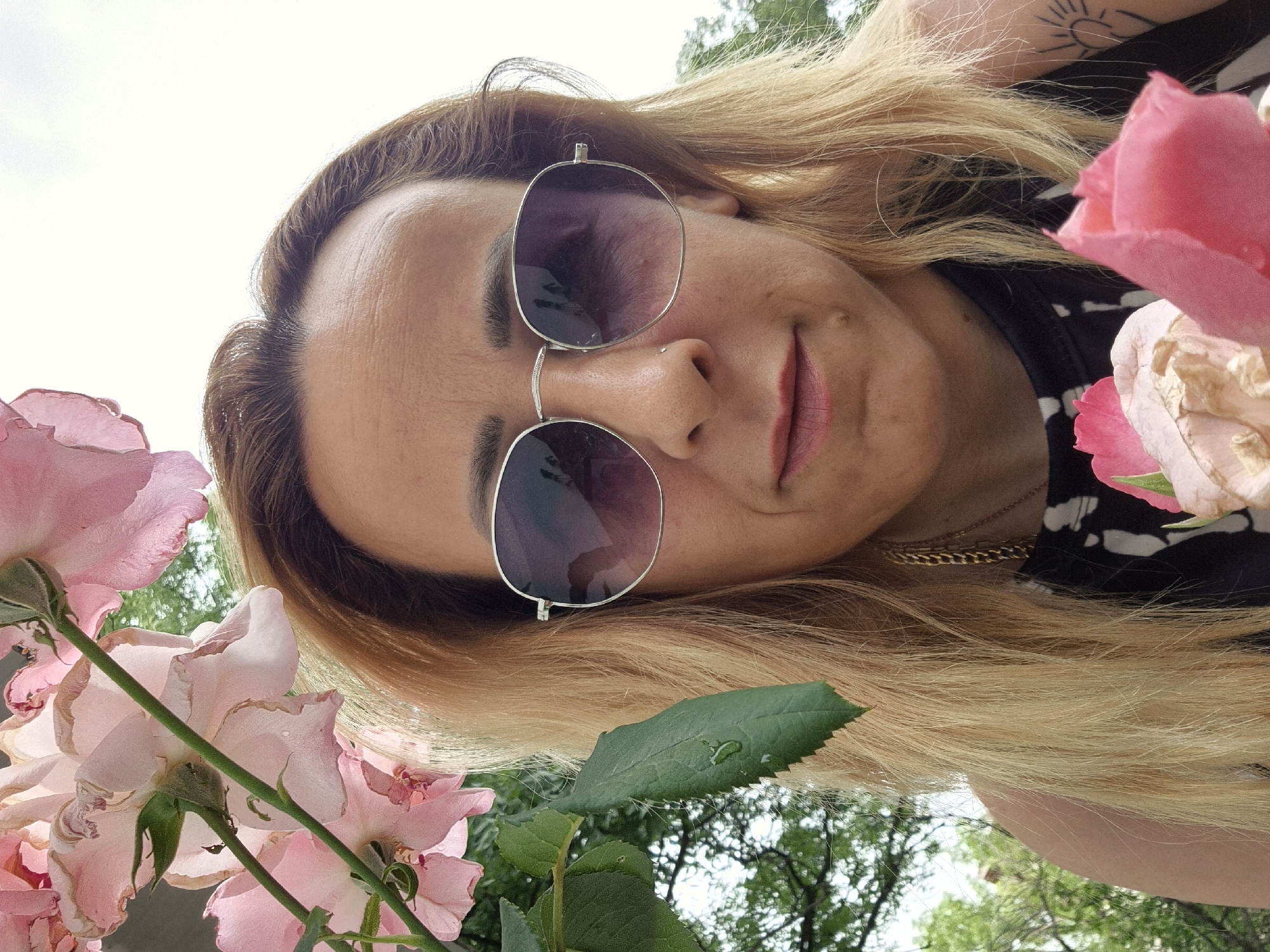 An individual, with fair skin and blonde hair, is photographed from the chest up, outdoors. They are wearing large, silver-rimmed sunglasses with gradient purple lenses, a gold chain necklace, and a black top with a white pattern. The individual has a nose ring and a tattoo on their left shoulder depicting a sun with radiating lines. They are holding two pink roses, one in front of their chest and one near their chin. Several pink rose blossoms and green leaves are visible in the foreground and on the left side of the image. The background consists of green foliage and a pale sky. The image appears to be a selfie taken in an outdoor setting, possibly a garden or park, during daylight.