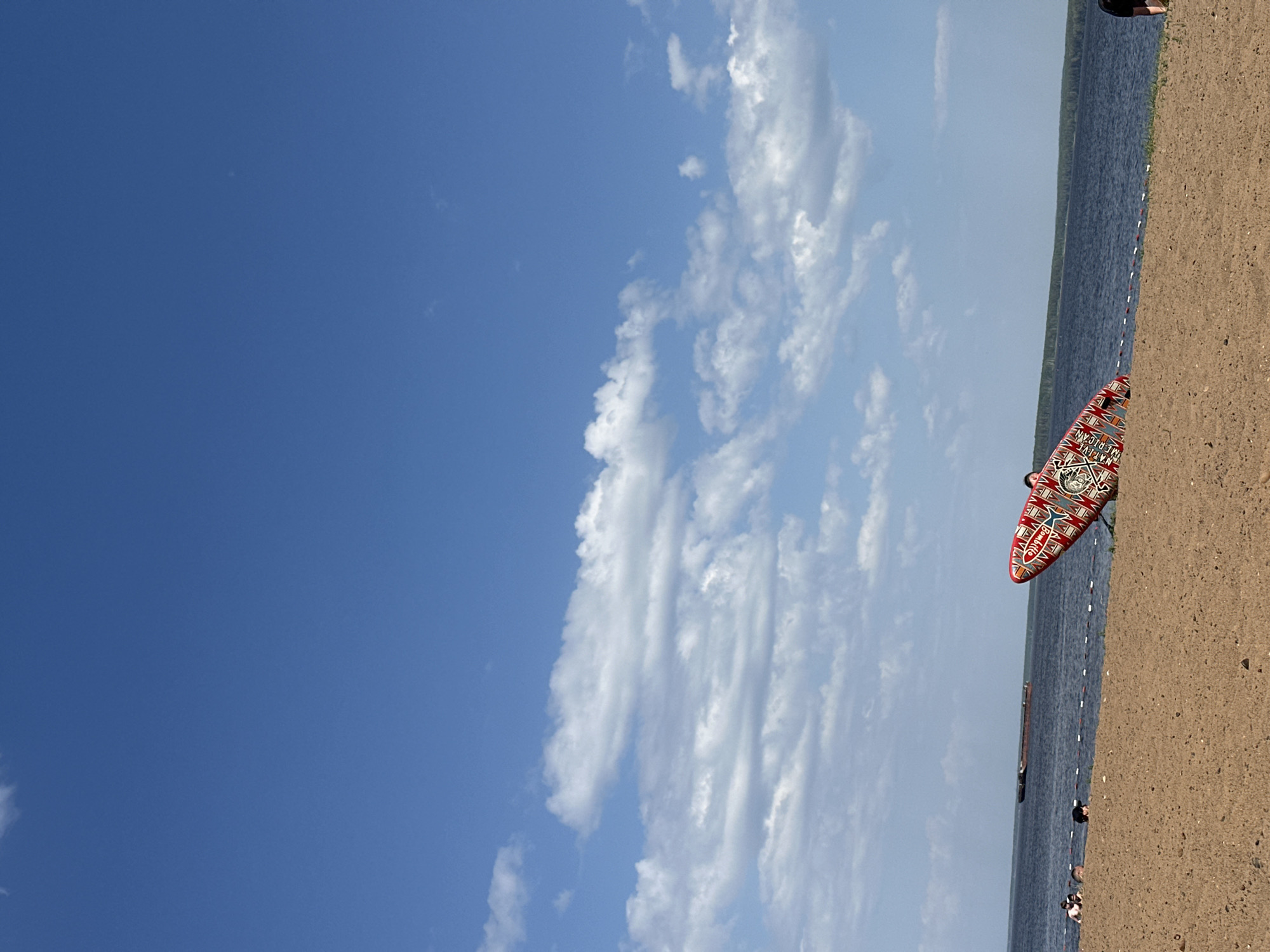 The image shows a beach scene in Chaikovsky, Russia. The sky is predominantly blue with scattered white clouds. A body of water is visible, running vertically in the frame, with a shoreline of sand. A decorated paddleboard is leaning against the sand near the water's edge. Several people are in the water. The light indicates it is daytime.