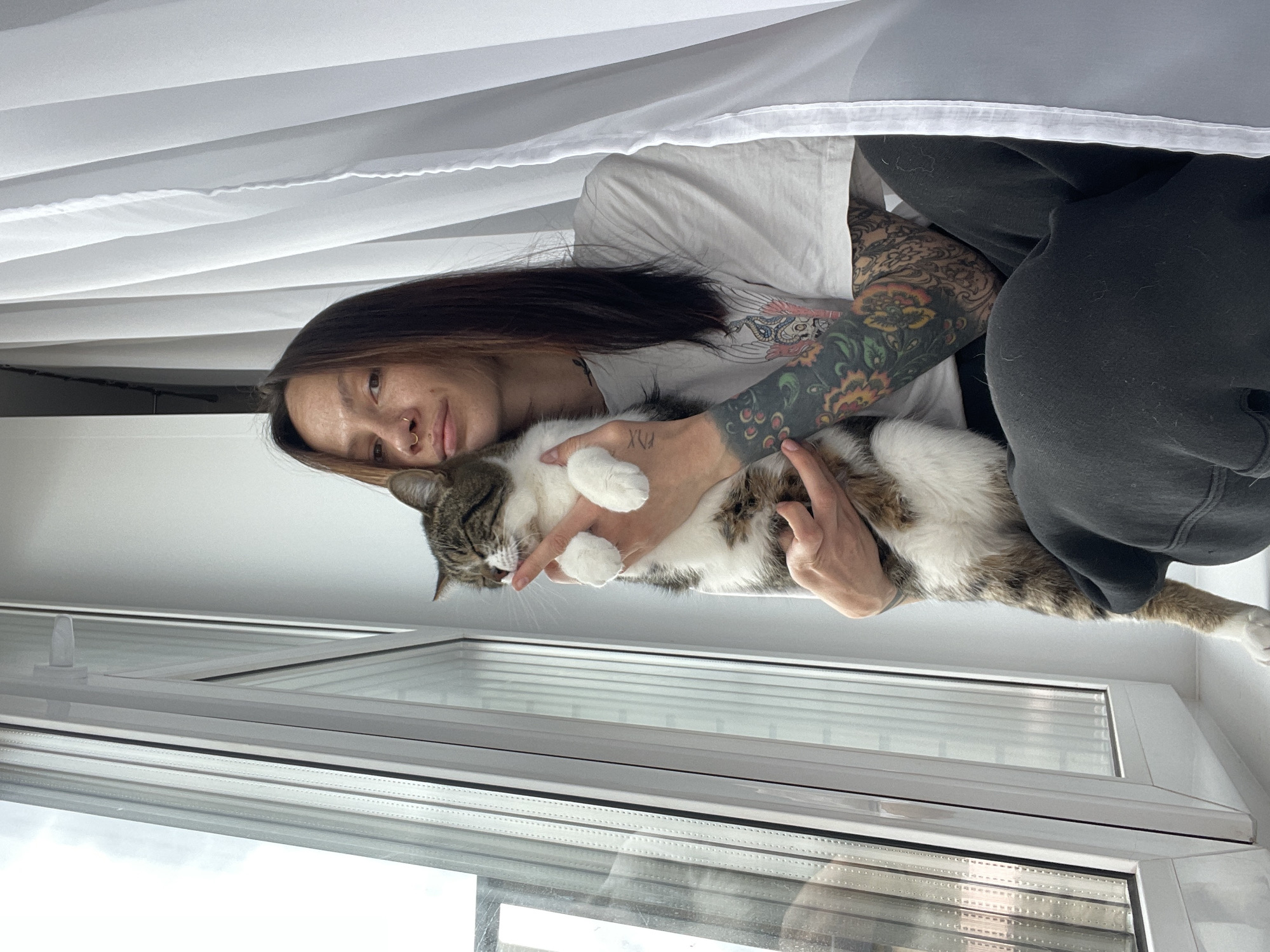 A woman with long dark hair and a nose ring sits in front of a white window in Pervomaysky, Russia. She wears a white T-shirt and dark pants. Her right arm is heavily tattooed. She holds a tabby and white cat, supporting its body with both hands. The cat's face is visible, and its paws are being held by the woman. A white curtain hangs above them. The window reflects the outside sky.