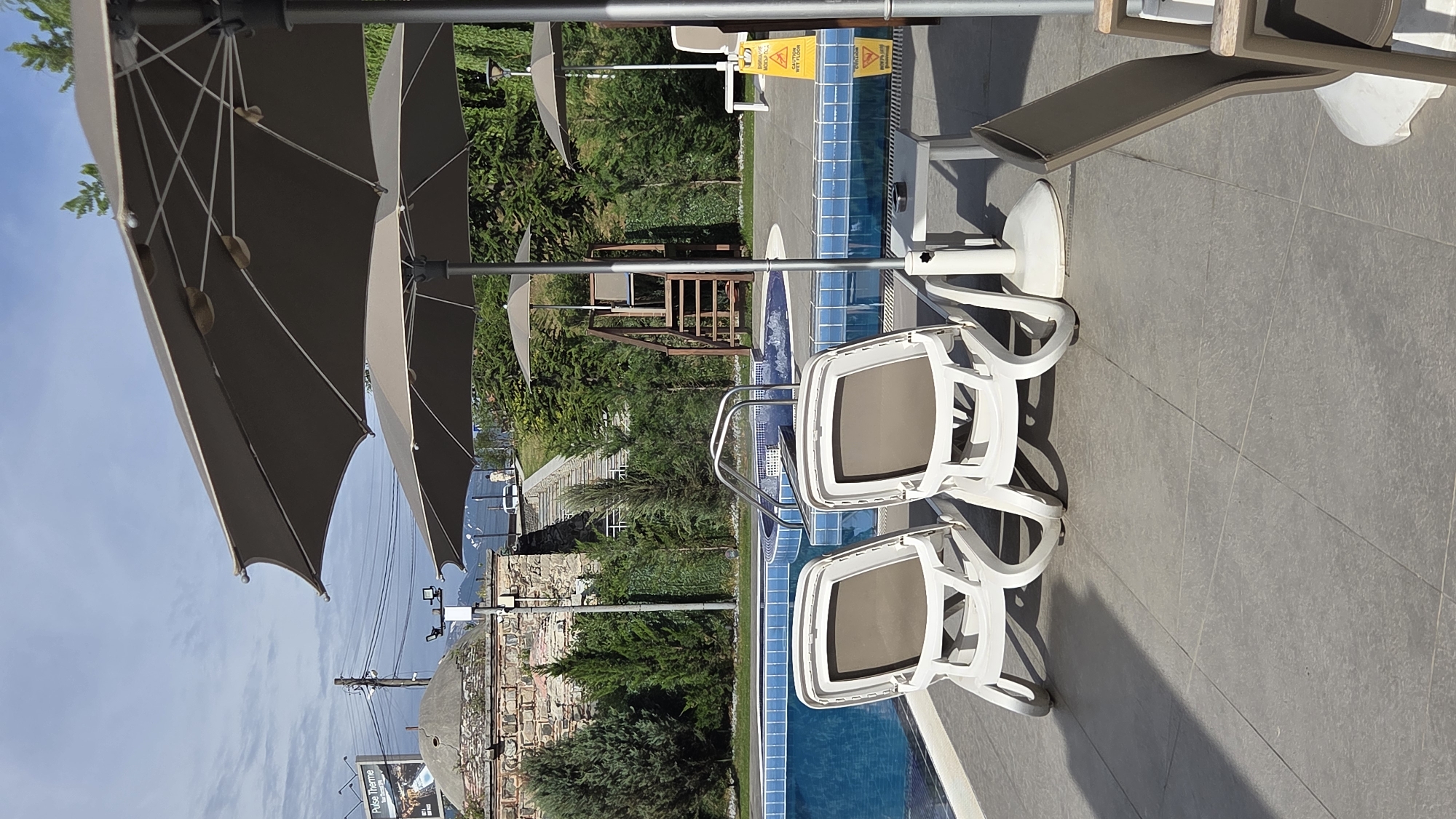 The image shows an outdoor swimming pool area on a sunny day. In the foreground, two white lounge chairs are positioned on a grey tiled patio. Behind the lounge chairs, a bright blue swimming pool with a jacuzzi section is visible. A set of metal stairs with handrails leads into the pool.
In the background, there is a wooden structure with a lifeguard chair. Further back, there are trees, a stone building with a domed roof, and a billboard with text that reads "Pulse Therme". A few power lines are also visible against the sky. The sky is partly cloudy. A yellow "Caution Wet Floor" sign is placed near the pool. Another chair and an umbrella are visible on the right side of the image.