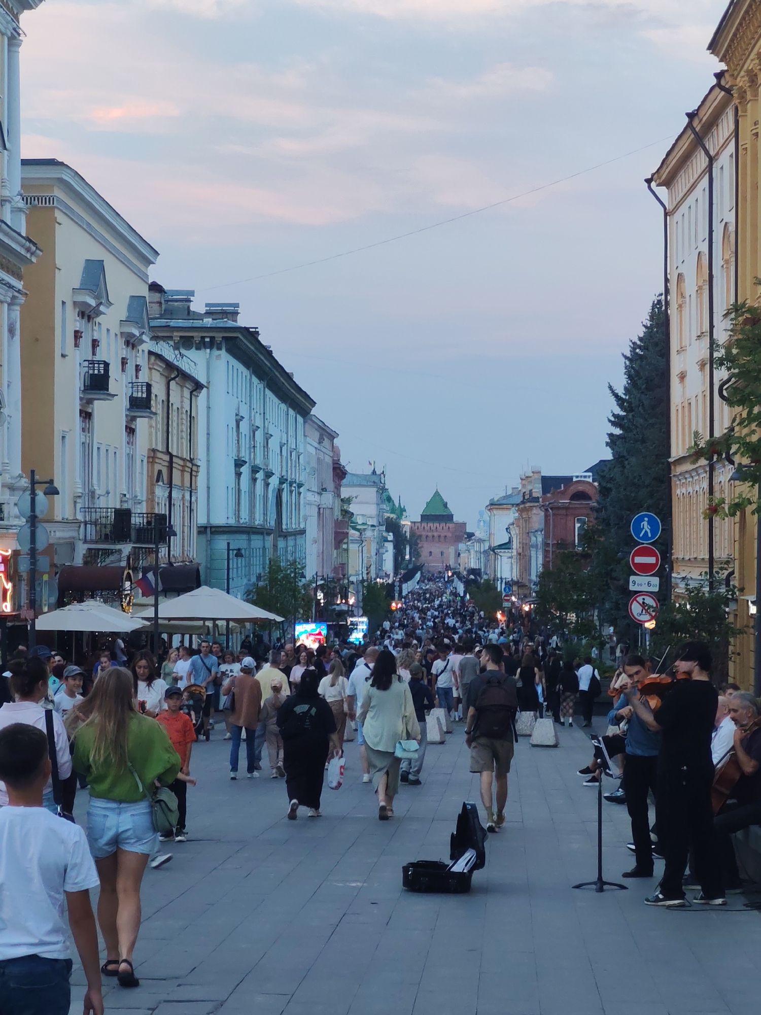 The image depicts a bustling pedestrian street in Nizhny Novgorod, Russia, likely during the evening hours, as indicated by the twilight sky. The street is lined with historic multi-story buildings with distinct architectural styles. A large crowd of people is walking along the cobblestone pavement, creating a lively atmosphere. On the right side of the street, a small group of musicians is performing, with a violinist and a cellist playing for passersby. Signs are visible, including traffic signs, suggesting the area is a public thoroughfare. The overall scene conveys a vibrant urban environment with cultural and social activity.