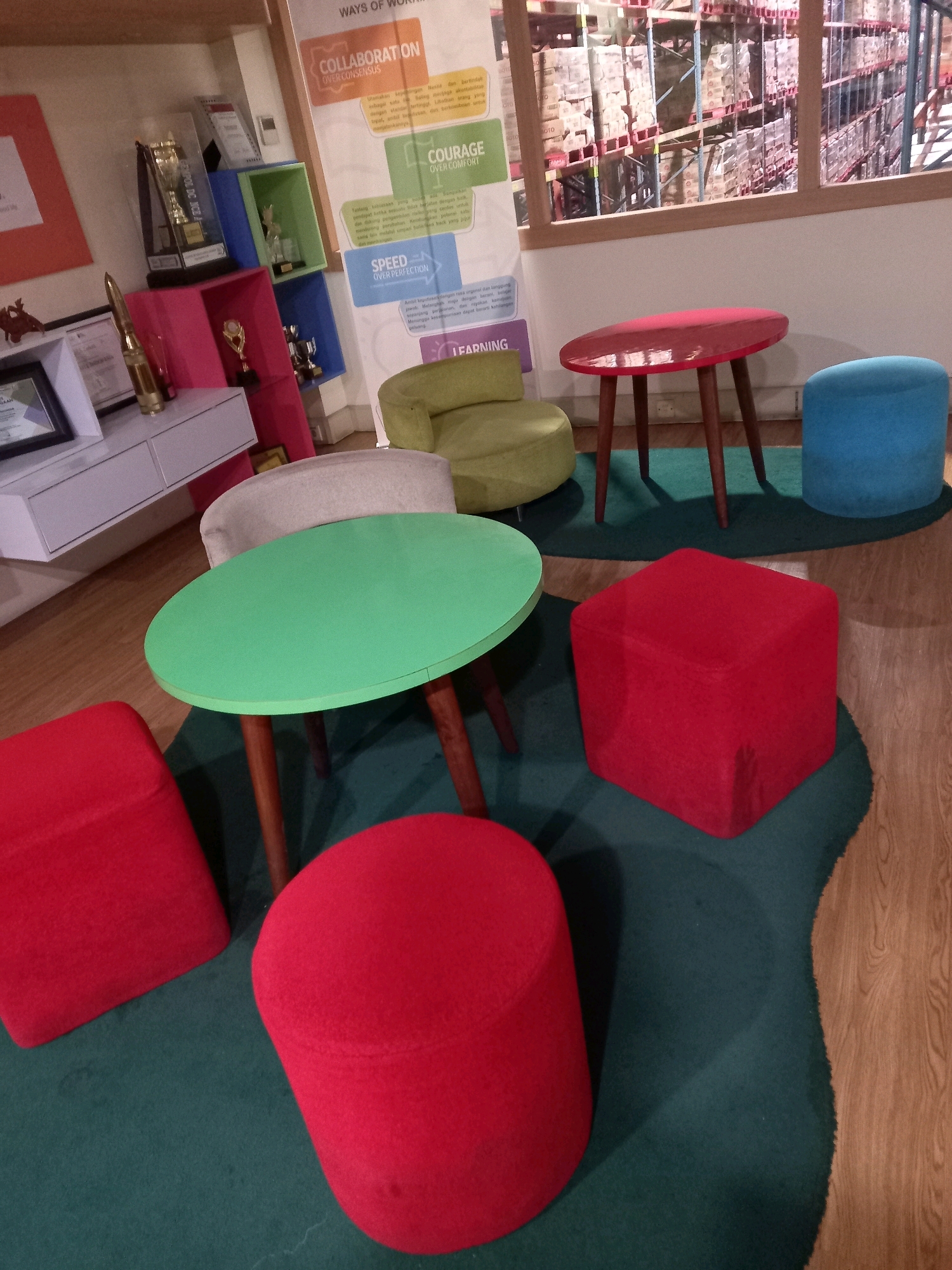 An indoor scene displays a collection of colorful, modern furniture arranged on a dark green rug over a wood-patterned floor. In the foreground, two bright red seating elements are visible: a cubic ottoman to the right and a rounded stool to the left. A round, mint green table with wooden legs stands in the center. Behind this, a beige upholstered chair is partially visible. Further back, a lime green, egg-shaped chair is situated near a bright red, oval-shaped table with wooden legs. To the right of the red table is a bright blue cylindrical ottoman. In the background, a wall features a motivational poster with sections titled "COLLABORATION," "COURAGE," "SPEED," and "LEARNING," alongside colorful shelving units displaying trophies and decorative items. A white wall-mounted cabinet with drawers is visible to the left, and a window in the upper right corner offers a view of a warehouse interior with stacked boxes on shelves. The lighting appears to be artificial and diffuse.