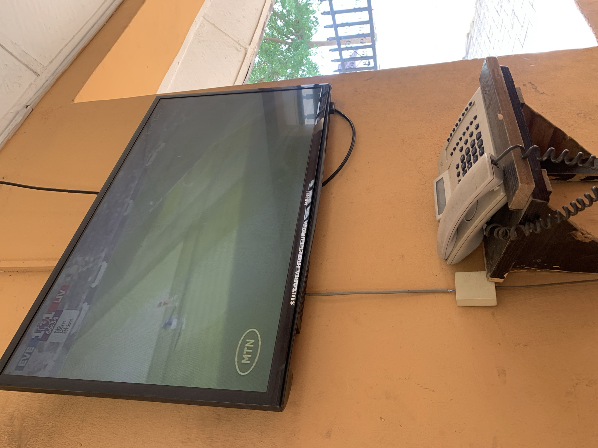 A wall in a building in Lokoja, Nigeria, has a flat-screen TV mounted on the wall, showing a soccer game with the MTN logo displayed. Next to the TV is a white office phone mounted on a wood bracket with a coiled phone cord. A gray cable runs along the wall beneath the TV and the phone. The wall is painted a light orange color. A section of a white ceiling is visible in the upper left corner of the image. A glimpse of a window and a metal railing are seen above the television.