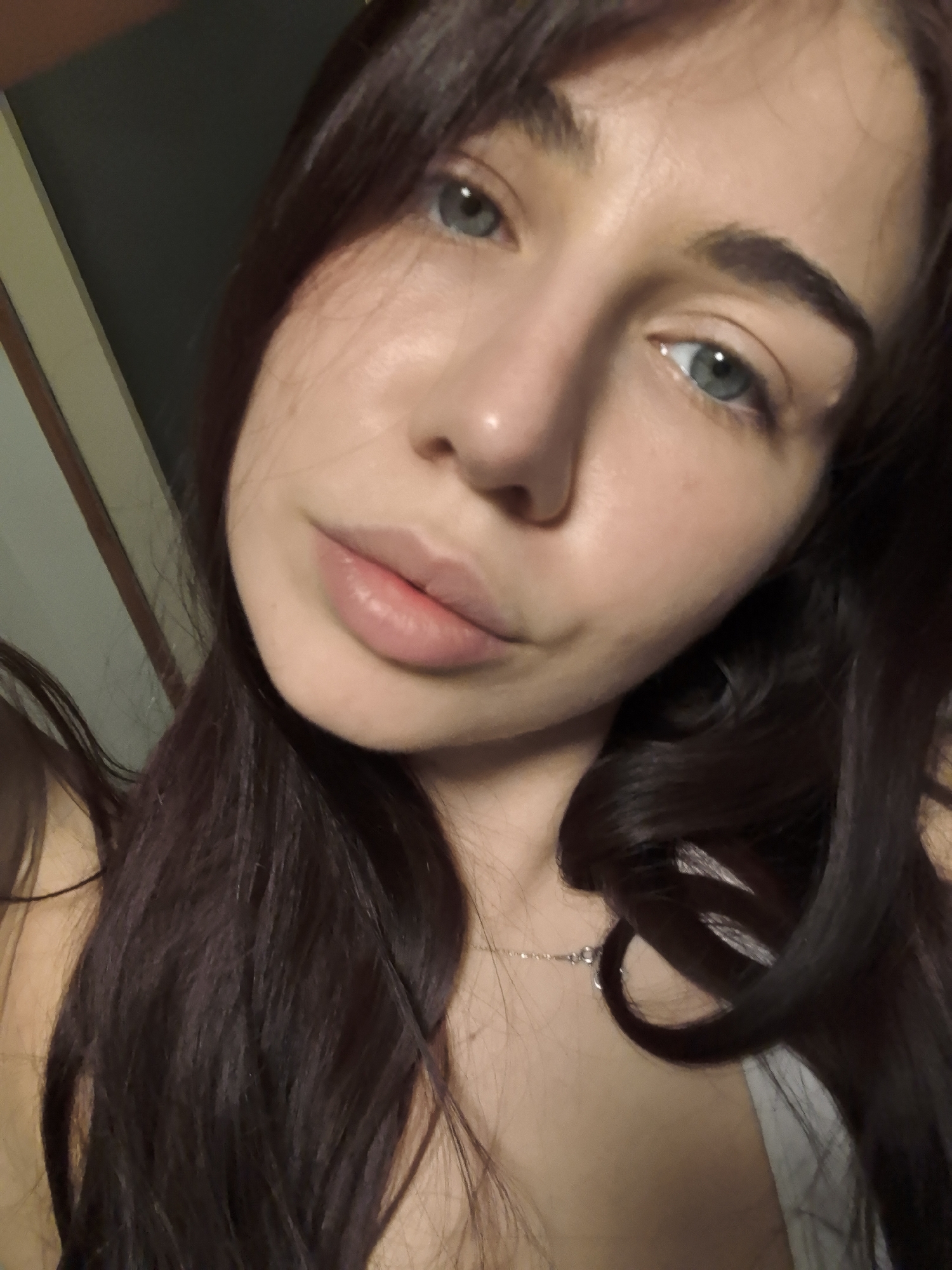 A close-up selfie depicts a young woman with fair skin, striking blue-green eyes, and full lips. She has dark brown hair, some strands of which fall across her face. She is wearing a delicate silver necklace. The background is indistinct, appearing to be an interior space. There is nothing indicating this image was taken in Stara Zagora, Bulgaria.