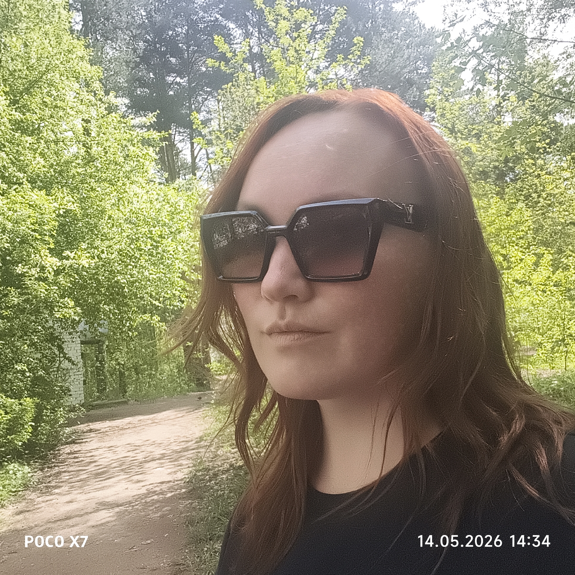 A woman with reddish-brown hair is wearing large, dark sunglasses. She is dressed in a black top. The background shows a dirt road and lush green trees, indicating an outdoor, possibly wooded area. The timestamp on the photo is "14.05.2026 14:34". The camera is a POCO X7.
Given the context of the city of Chaikovsky, Russia, the location may be a park or natural area within or near the city. However, it is not possible to know the specific location.