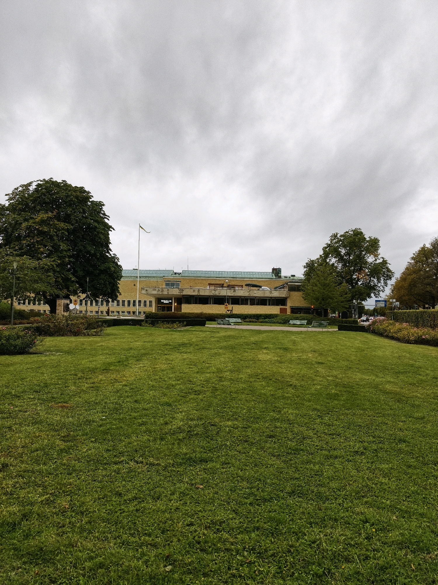 The image depicts the Östergötlands museum in Linköping, Sweden. A large, well-maintained lawn occupies the foreground of the scene. In the middle ground, a long, low-rise building with a yellow facade is visible, featuring numerous windows and a prominent entrance. A flagpole with a yellow and green flag stands to the left of the building. Several green benches are placed on a paved area in front of the building, and manicured hedges line the front. Large trees are situated on either side of the building, one on the left being a mature, leafy specimen, and another on the right. The sky above is overcast, with a dense layer of grey clouds. A few vehicles are visible on a street to the right of the museum. The overall atmosphere is that of a public park or civic space under a cloudy sky.