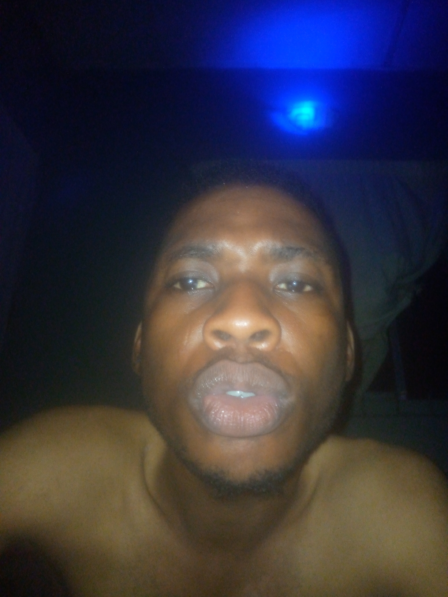 A close-up, low-light portrait features a person, likely male, with dark skin, directly facing the camera. The shot captures the individual from the chest up, with their shoulders and a portion of their bare chest visible, suggesting they are shirtless or wearing minimal clothing. Their eyes are open and looking forward, and their lips are slightly parted. Short, dark hair is visible on their head, and faint stubble can be seen on their chin.
The setting appears to be a very dim indoor space or nighttime, as the background is predominantly dark black. A distinct, bright blue light source is situated in the upper center of the frame, casting a blue ambient light, especially on the upper parts of the image and subtly on the subject's skin. This artificial illumination creates a moody atmosphere. The person is likely taking a selfie, given the close proximity and direct gaze. No other objects or text are visible in the image. The expression of the person is neutral.