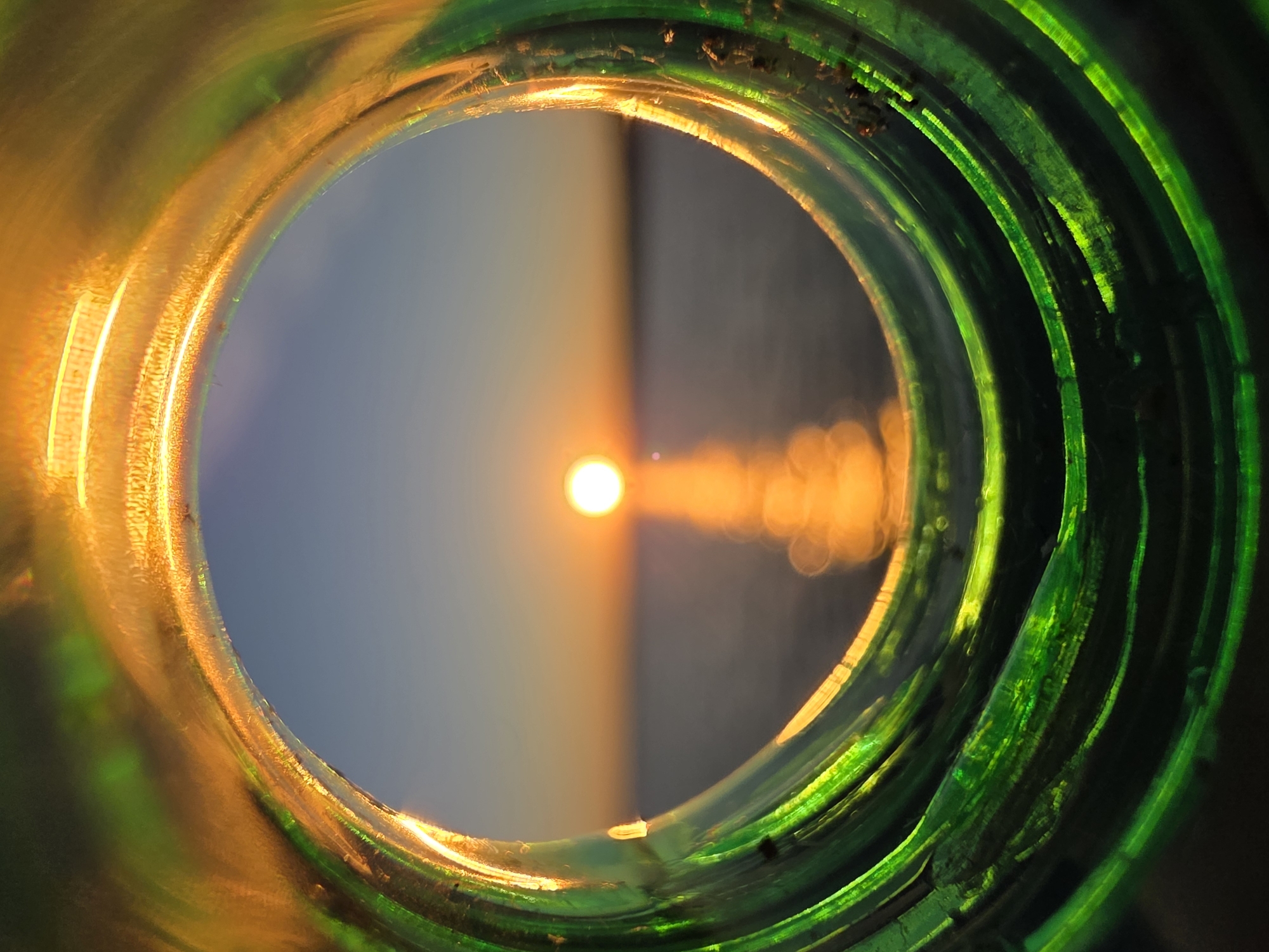 The image is a close-up shot looking through the opening of a green glass bottle. The bottle's rim is textured and catches the light, creating a halo effect with streaks of green and golden hues. Beyond the bottle's opening, a sunset is visible over a body of water, possibly the Kama River, given the implied location in Chaikovsky, Russia. The sun, a bright orange orb, is low on the horizon, casting a shimmering reflection on the water. The sky transitions from a pale blue near the horizon to a darker hue above. The overall impression is a framing of a natural scene through an artificial object.