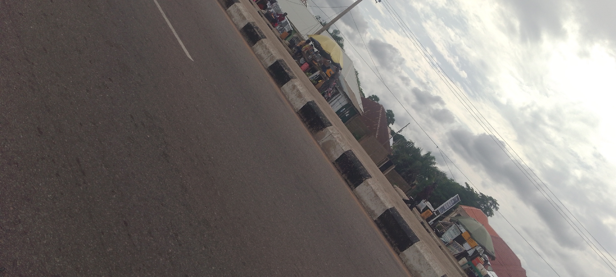 The image shows a street scene in Jalingo, Nigeria, under a cloudy sky. A road with a white line marking is visible in the foreground, bordered by a curb with alternating black and white painted sections. Along the curb, a market appears to be set up with various stalls and vendors. A yellow umbrella is open, providing shade over one of the stalls. Several people are visible, some appearing to be vendors and others customers, engaged in what seems to be a transaction. Buildings and trees are present in the background, suggesting an urban or semi-urban environment. Overhead power lines are also visible crisscrossing the sky. The overall impression is of a busy marketplace situated alongside a road.