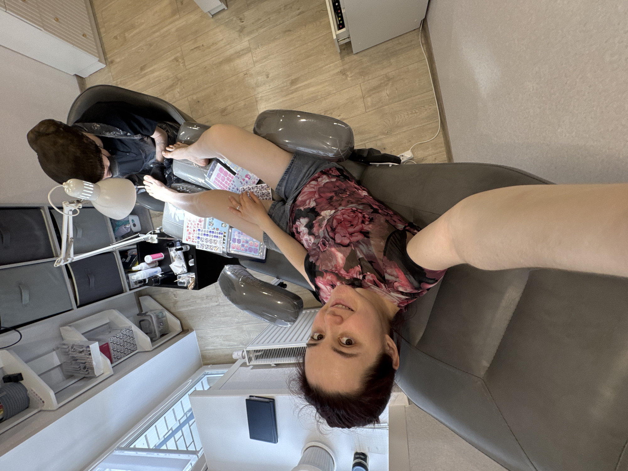 The image is taken from a high angle, looking down at a woman in a nail salon, possibly in Izhevsk, Russia. The woman is sitting in a large, grey chair with her legs extended towards a person in a pedicure chair. The person in the pedicure chair is obscured by a dark covering. The woman in the large chair is wearing a floral top and shorts, and she is looking at the camera with a slight smile. In front of her are nail stickers and other nail art supplies on a table. Next to the table is a white shelving unit with storage boxes and supplies. A lamp is positioned to illuminate the work area. A radiator and an air purifier are also visible in the background. The overall impression is of a relaxed and well-lit nail salon setting.