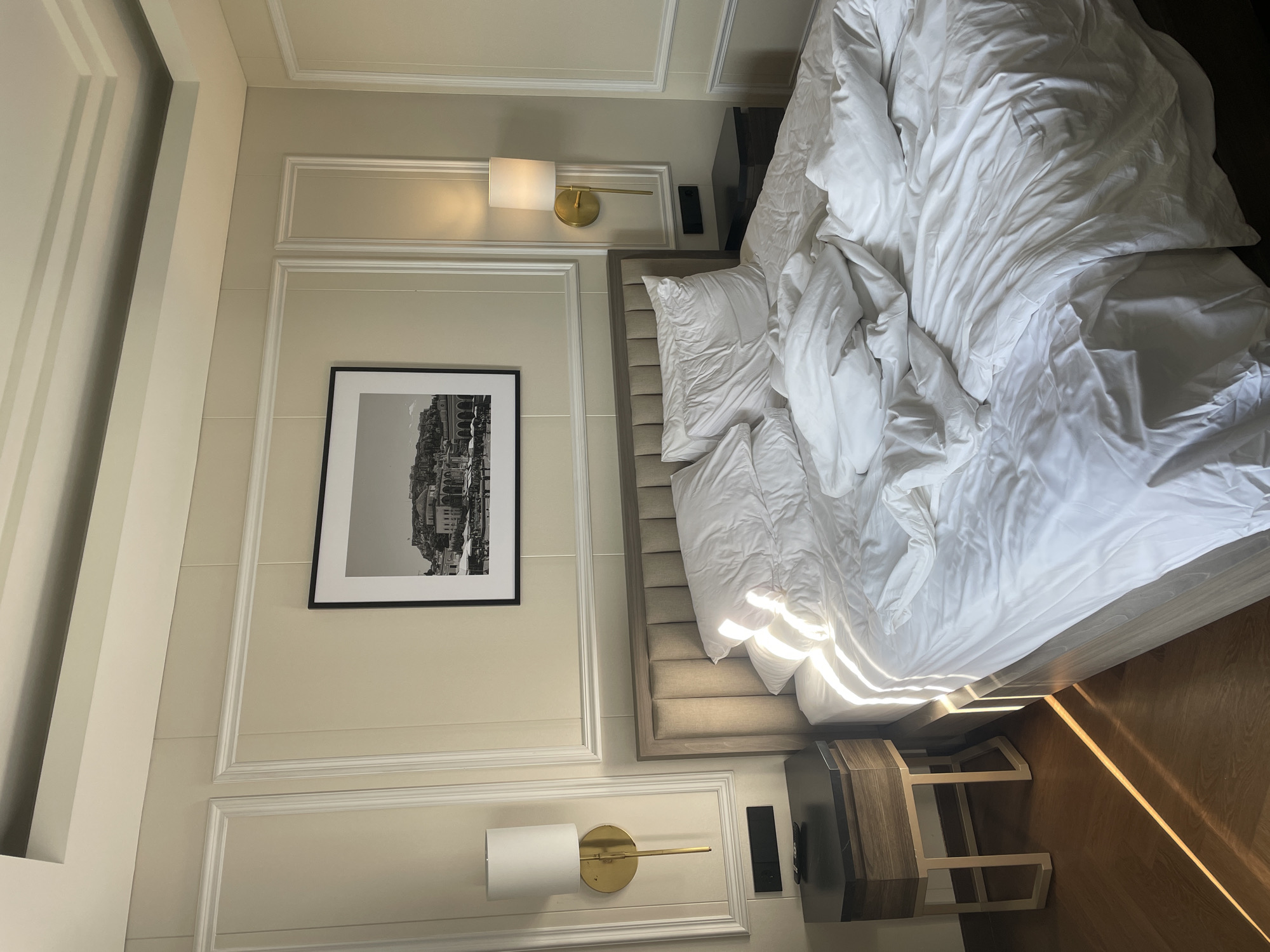 This image captures a wide shot of an unmade bedroom, likely in a hotel or modern home, bathed in natural sunlight. The central focus is a large bed covered in rumpled white linens, including a duvet and several pillows, indicating recent use. The bed features a light beige, upholstered headboard with vertical stitching, and a matching wooden bed frame.
On either side of the bed, there are modern, dark-topped hexagonal bedside tables with wooden bases and light-colored, geometric metal legs. Above the left bedside table, a black electronic control panel is mounted on the wall. Wall-mounted sconces with cylindrical white shades and long, slender gold-toned arms are positioned on both sides of the bed; the one on the right appears to be illuminated.
The walls are painted a light cream or beige color and feature elegant white decorative molding, creating a paneled effect. Centered above the bed, a large black-framed black and white photograph depicts an urban landscape, possibly a European city with buildings on a hillside overlooking a body of water and a waterfront promenade. The ceiling is recessed and multi-layered, also in a light color, adding architectural detail.
Strong natural sunlight streams into the room from an unseen window to the left, casting distinct, diagonal stripes of light and shadow across the white bedding and extending onto the warm, reddish-brown wooden floor. The overall ambiance is bright and serene. There are no people present in the scene.
