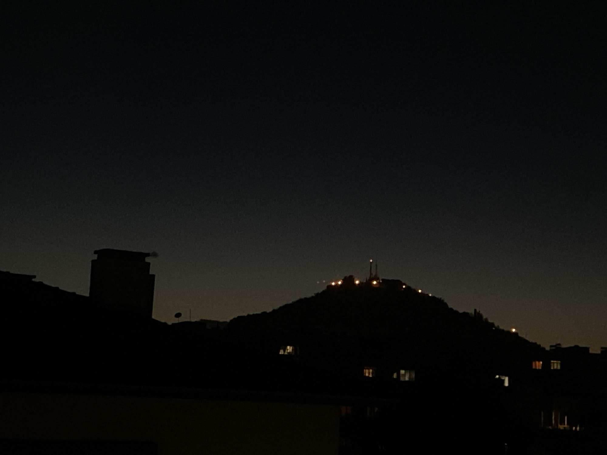 The image captures a nighttime silhouette of a city landscape. In the foreground, the dark shape of a building with a prominent chimney dominates the left side. To the right, a larger hill or mountain is visible, topped with illuminated structures, possibly a communication tower or antenna array, along with scattered lights that suggest buildings or settlements on its slopes. The sky is a gradient of dark blue to a lighter, warmer hue near the horizon, indicative of twilight or distant city glow. Several windows with interior lights are visible on buildings nestled at the base of the hill. The overall impression is one of urban sprawl against a darkening sky.