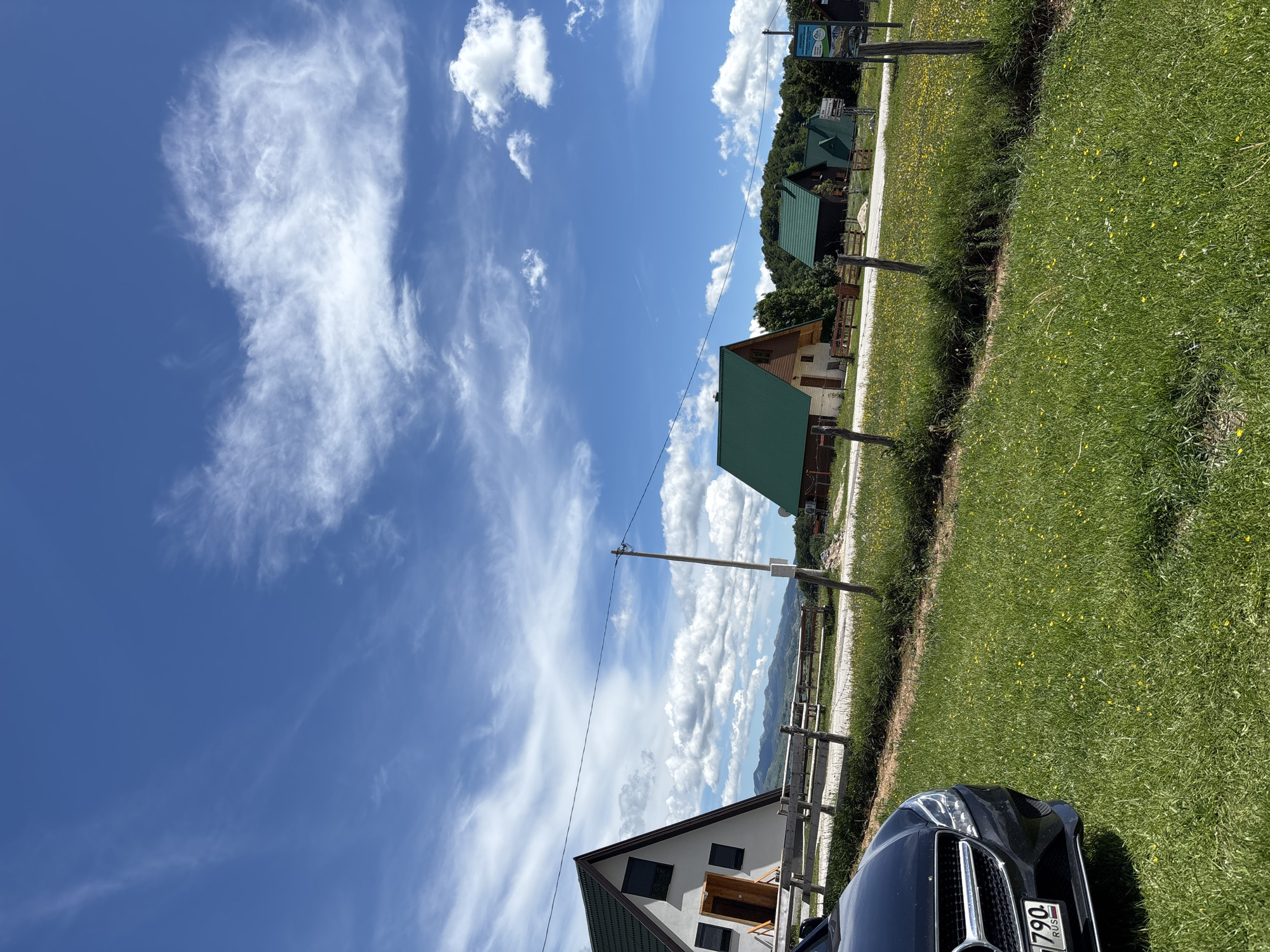 In the city of Virak, Montenegro, on a bright, clear day with scattered white clouds in a blue sky, a black Mercedes-Benz sedan is parked on a grassy area. The car is angled towards the left side of the frame, showing its front bumper and grille. Behind the car, a wooden fence runs horizontally across the scene, separating the grassy foreground from a gravel path and a row of houses. These houses are characterized by their steep, green-pitched roofs and are nestled amongst trees on a gentle slope. In the background, a range of mountains is visible under the expansive sky. The environment suggests a rural or semi-rural setting, possibly a village or resort area. There are no people visible in the image.