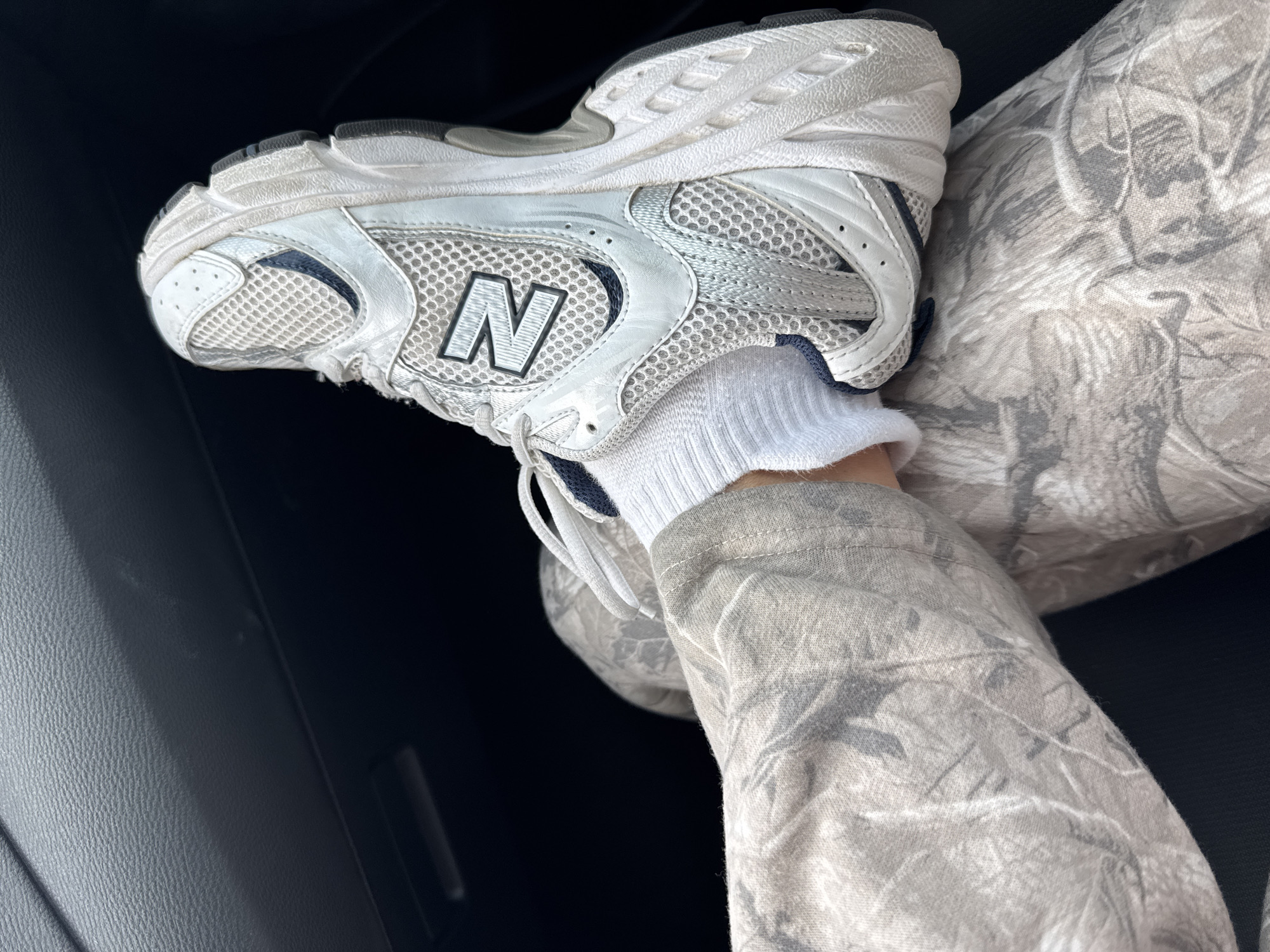 A close-up shot depicts a leg wearing a white athletic shoe with a prominent black "N" logo, white socks, and camouflage pants. The leg is positioned against a dark background, possibly the interior of a vehicle. The setting is not identifiable as Riverside, California, or any other specific location.