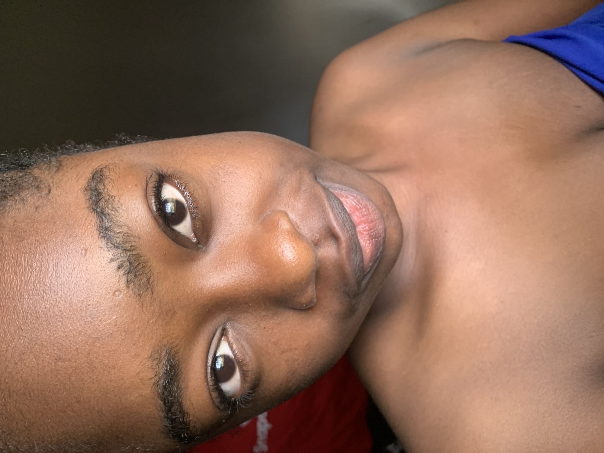 A close-up, head and shoulders portrait of a young Black woman is presented, likely taken indoors with natural light. The woman has dark skin, dark hair, and dark eyes. She is looking directly at the camera with a slight smile. Her shoulders are bare. A hint of a blue garment is visible at the bottom right of the frame. The background is dark and out of focus. The image does not contain any information about the location in Rafin Par, Nigeria.