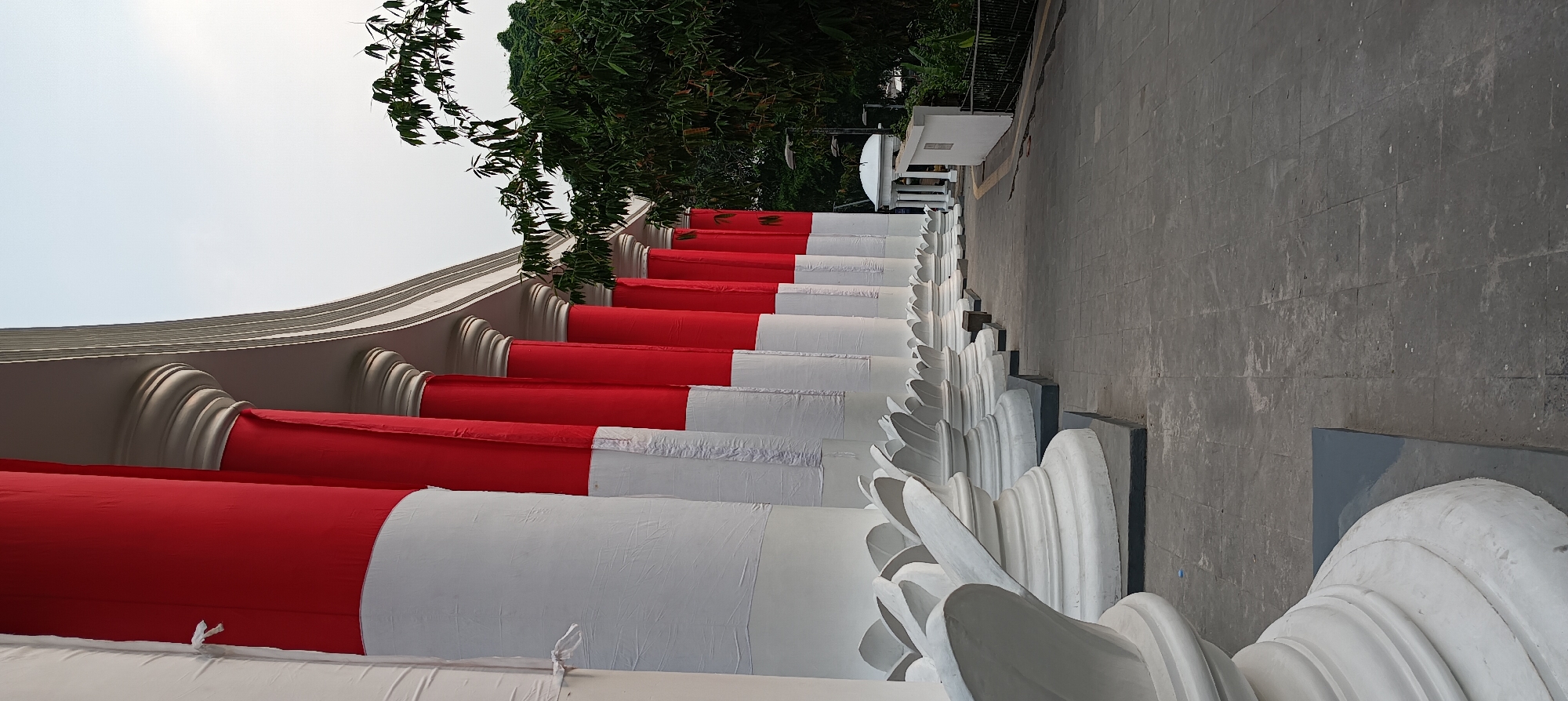 The image shows a low-angle view of the exterior of a building with large, decorative pillars. The pillars are wrapped in red and white fabric, resembling the Indonesian flag. They stand in a row, receding into the distance. The ground in front of the pillars is paved with grey tiles. To the right of the pillars, there is a pathway leading to a white gazebo-like structure. Trees and foliage are visible in the background. The sky is overcast. This scene is likely the exterior of the Bogor Botanical Gardens, a popular tourist attraction in Bogor, Indonesia, known for its classical architecture and lush greenery.