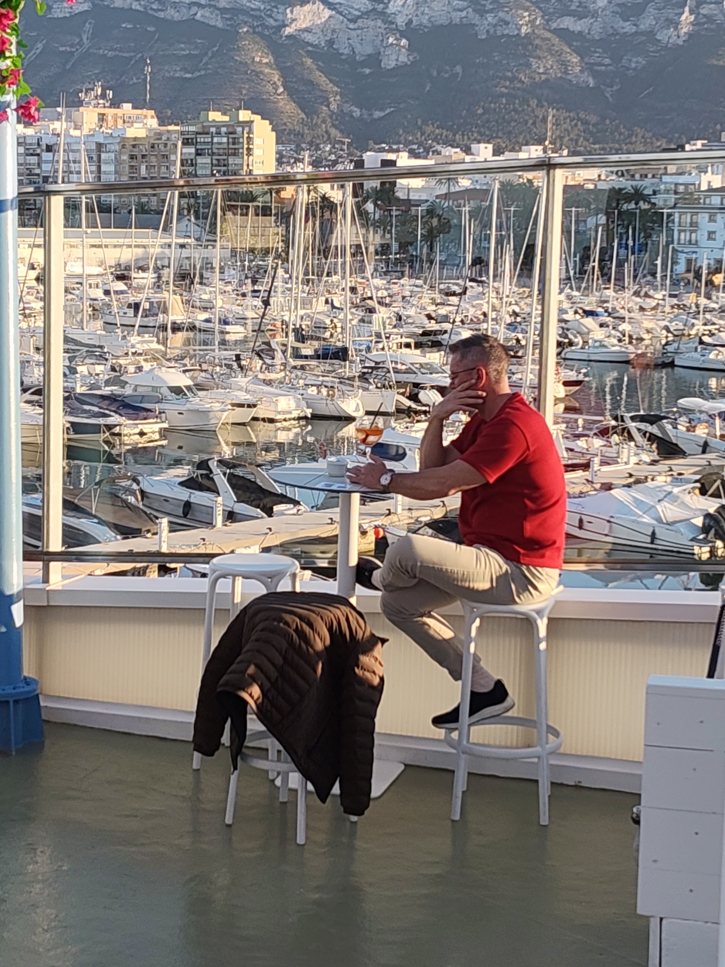 A middle-aged man with short hair and glasses sits at a small round table on a white high-backed stool, looking pensively out at a harbor filled with many sailboats and motorboats. He is wearing a red t-shirt, tan pants, and black shoes. He rests his right elbow on the table and his hand supports his face. A drink and a wristwatch are on the table. A brown jacket is draped over an adjacent stool. The location is the Marina de Dénia in Dénia, Spain, identifiable by the numerous boats and the building and mountainous backdrop.