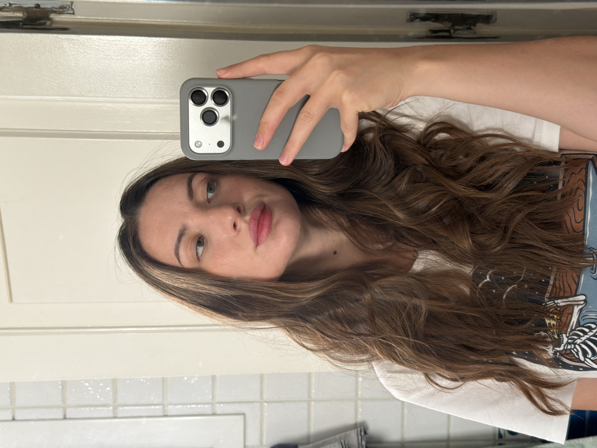 A young Caucasian woman with long, wavy brown hair is taking a mirror selfie. She is wearing a white t-shirt with a graphic design. She is holding a gray iPhone in her right hand, which is positioned to capture the reflection in a mirror. Her lips are slightly pursed, and she is looking at her reflection. The background appears to be a bathroom with white tiled walls and a closed door. There is no indication of the location being Sharm El Sheikh, Egypt.