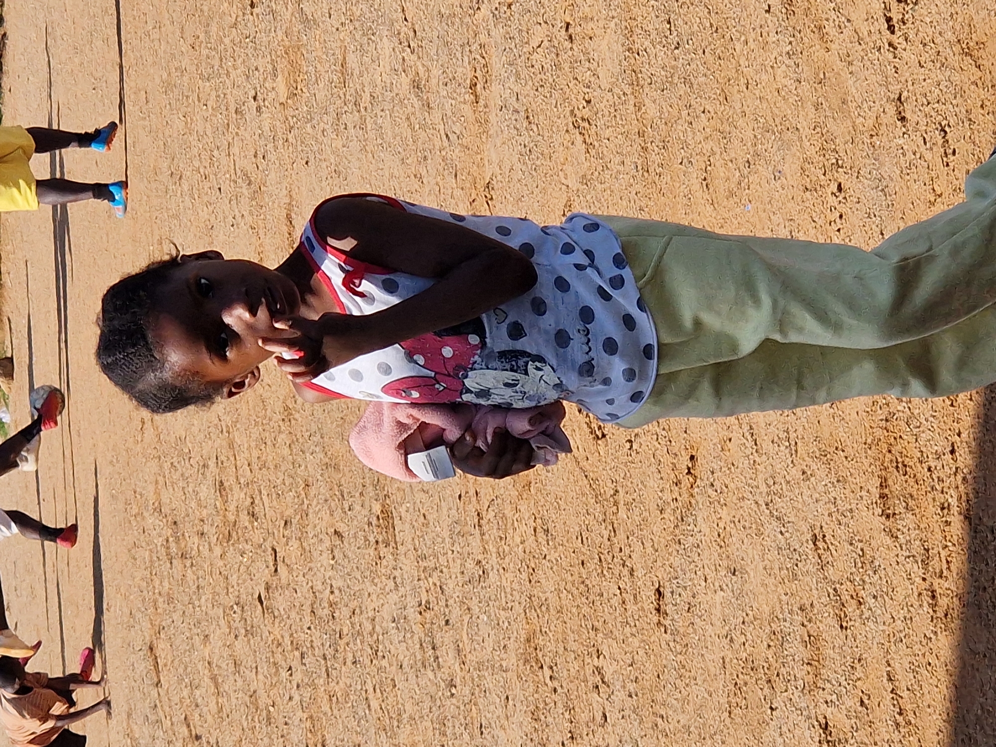 A young Black girl stands in the foreground, looking towards the camera with her right hand near her mouth. She is wearing a white sleeveless shirt with black polka dots and red trim, featuring images of Minnie Mouse. She also wears light green pants and carries a folded pink cloth in her left arm. In the background, two children are playing soccer on a sandy, dirt field. One child, wearing a light brown shirt and red shoes, is crouched down. Another child, in yellow shorts and blue soccer cleats, kicks a ball. The environment appears to be an outdoor area in Kabong, Nigeria, characterized by the sandy terrain and sparse vegetation.
